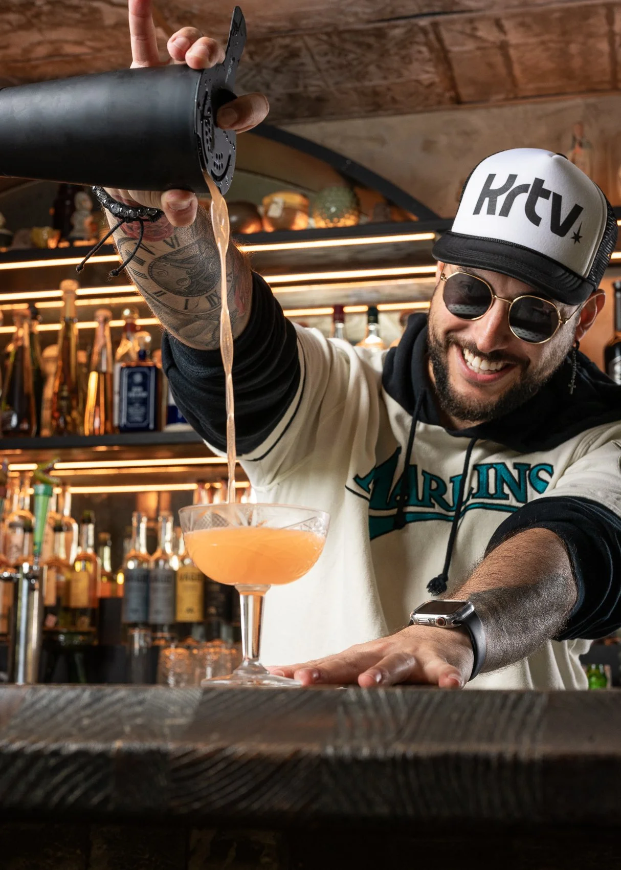 Bartender Stefano smiling while pouring a Naked and Famous cocktail at the mezcaleria bar inside Bodega Taqueria y Tequila in Coconut Grove, wearing a Miami Marlins jersey and sunglasses.