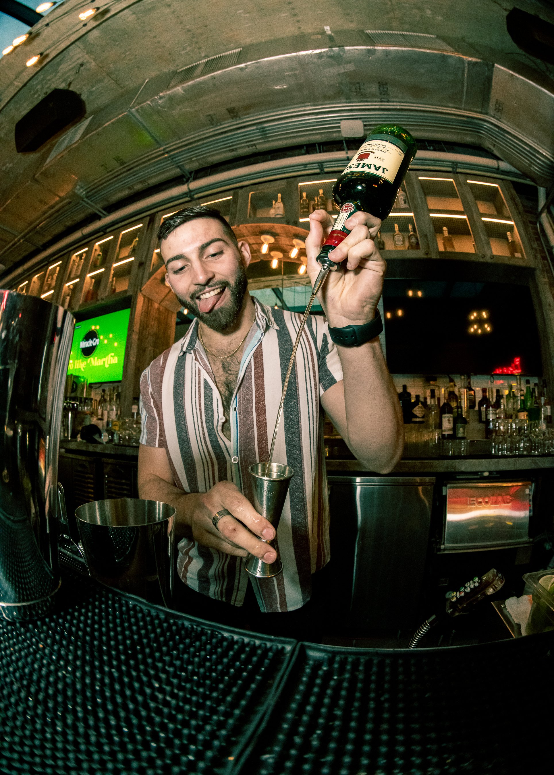 Bartender Alejandro Gil smiling as he measures and pours Jameson Irish Whiskey into a jigger at Bodega Taqueria y Tequila in Coconut Grove, captured behind the bar for his social media content.