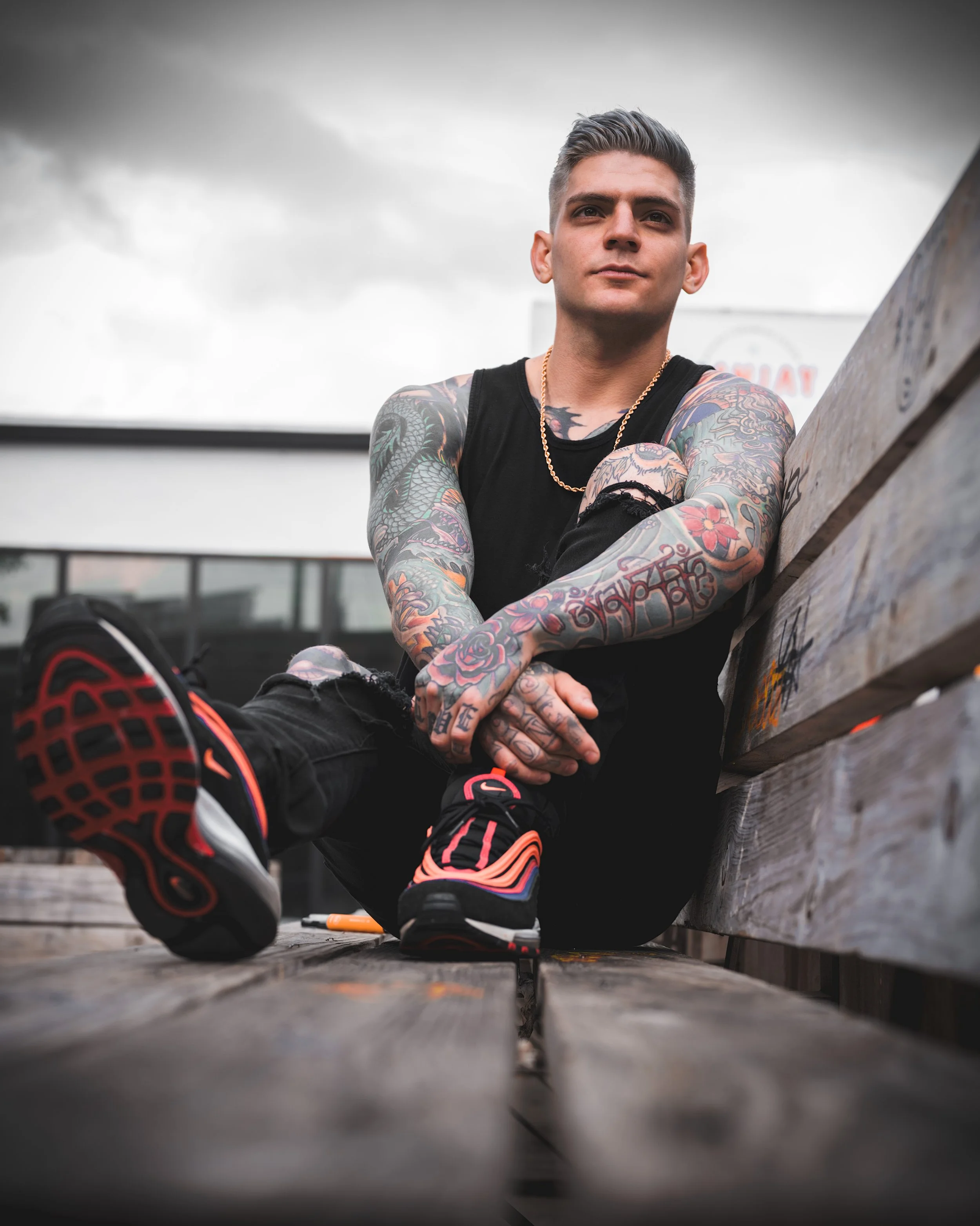 Outdoor portrait of Ramon Meneses sitting on a wooden bench, showcasing his tattooed arms and casual streetwear style, photographed for portfolio work.
