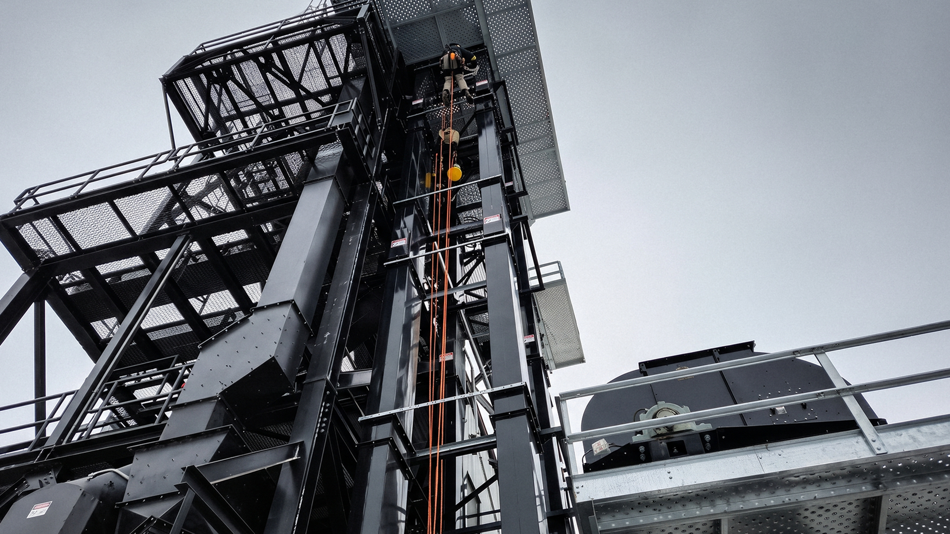 Low-angle view of a black steel grain elevator leg and headframe, with an APEX rope access technician working on ropes near the top.