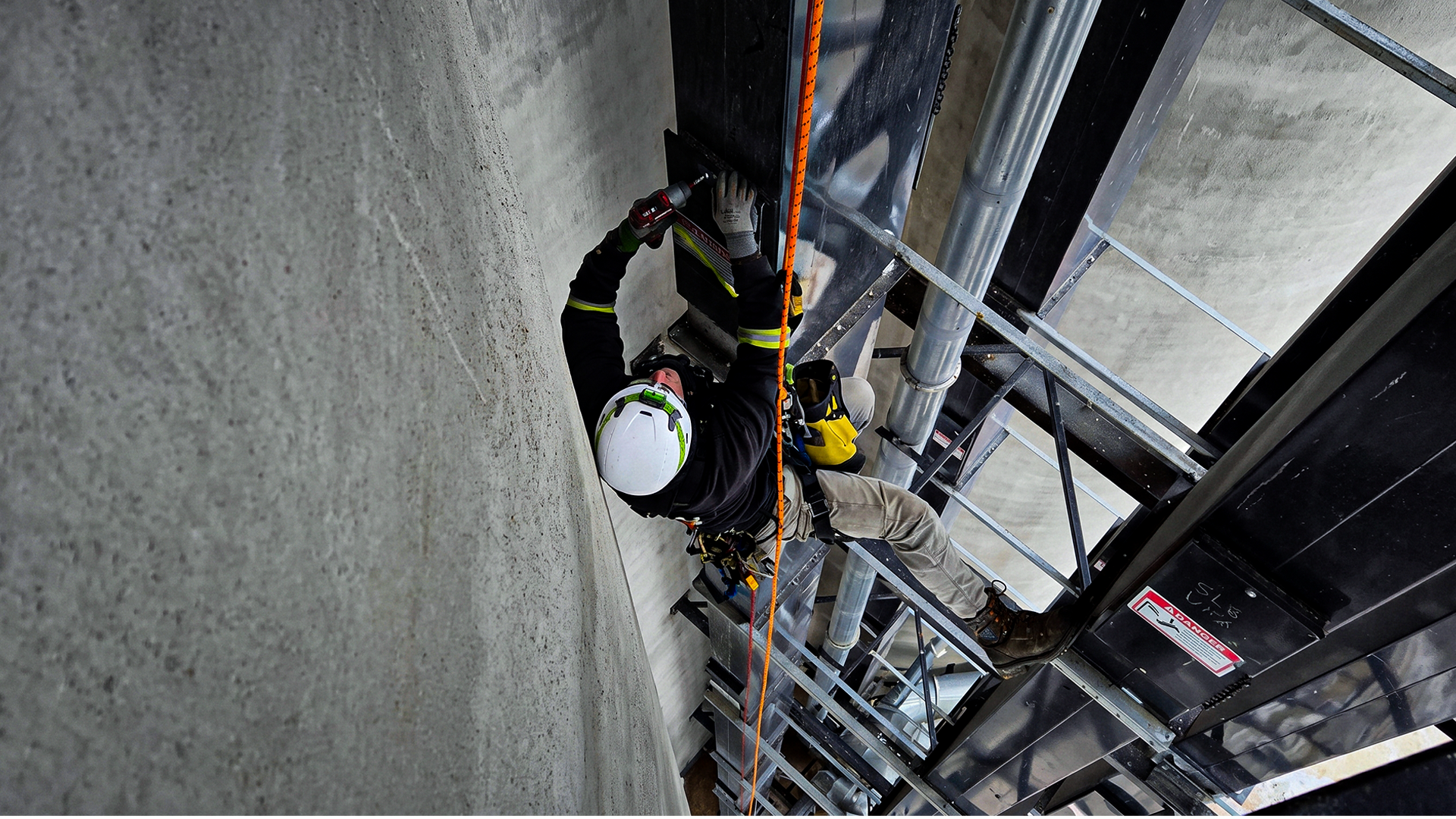 APEX rope access technician in a white helmet drives a replacement fastener into an explosion relief panel on a grain elevator, working from rope.