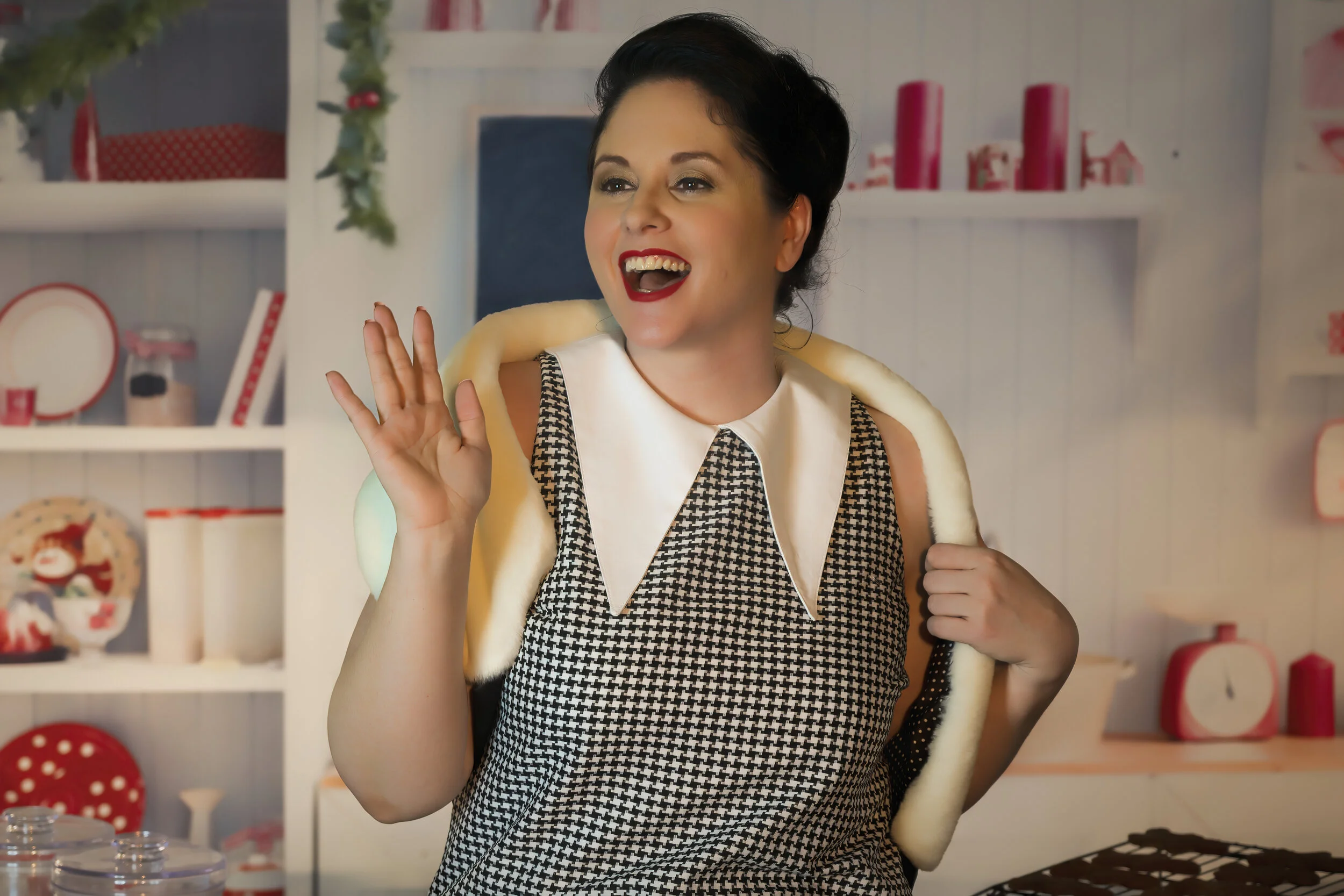 A woman with short dark hair, red lipstick, and a checked dress waving and smiling in a festive kitchen decorated for Christmas.