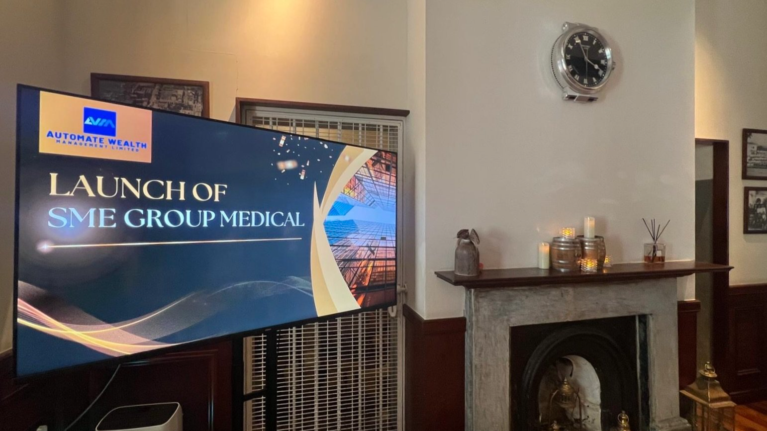 TV screen displaying a presentation titled 'Launch of SME Group Medical' with a logo of Automate Wealth Management Limited in the top left corner, set in a room with a mantle decorated with candles, a bottle, and a diffuser, and a wall clock above.