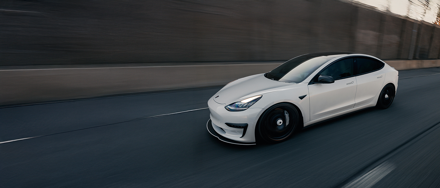 A white Tesla Model 3 driving on a highway at dusk with a concrete barrier on the side.