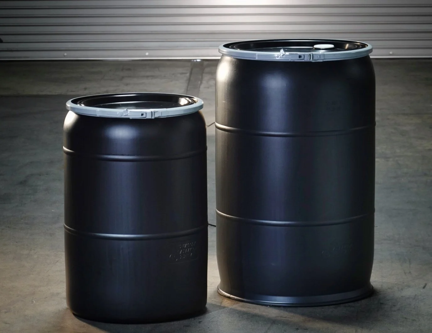 Two black metal drums, one smaller and one larger, are standing on a concrete floor in front of a metal roll-up door.