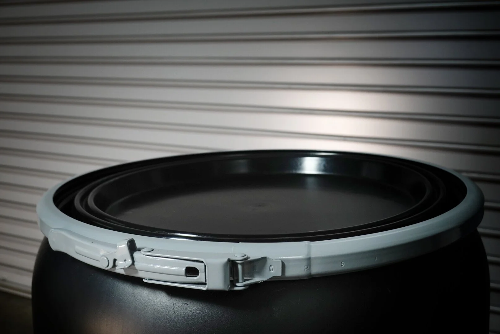 A black trash can with its lid partially closed, set against a gray corrugated metal background.