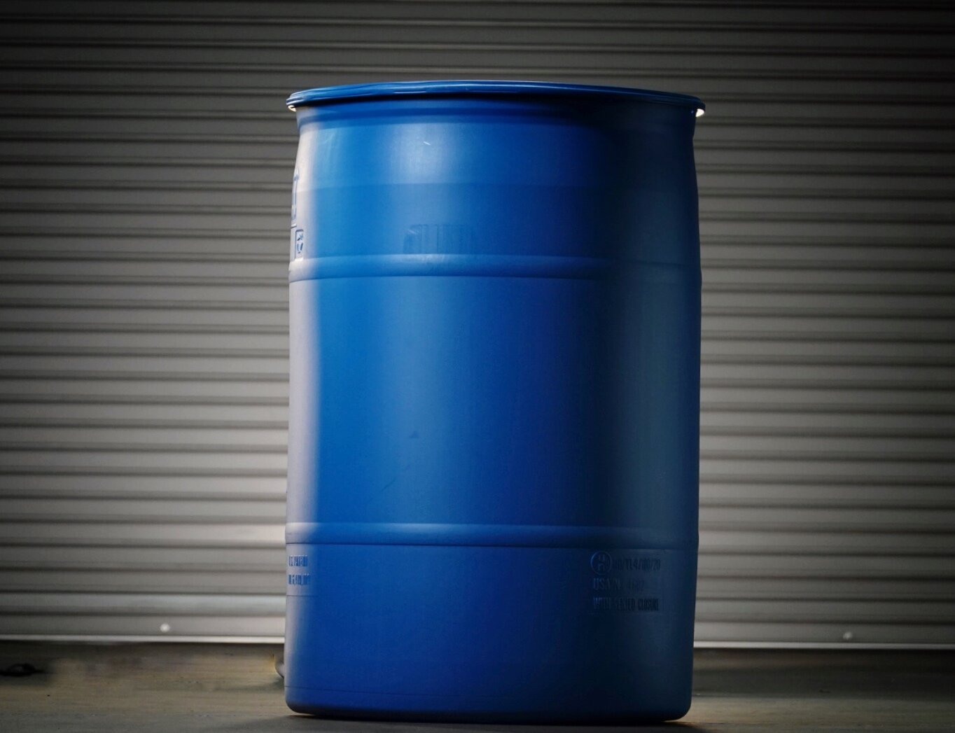 A blue industrial barrel standing in front of a metallic roll-up door, illuminated by light from the side.