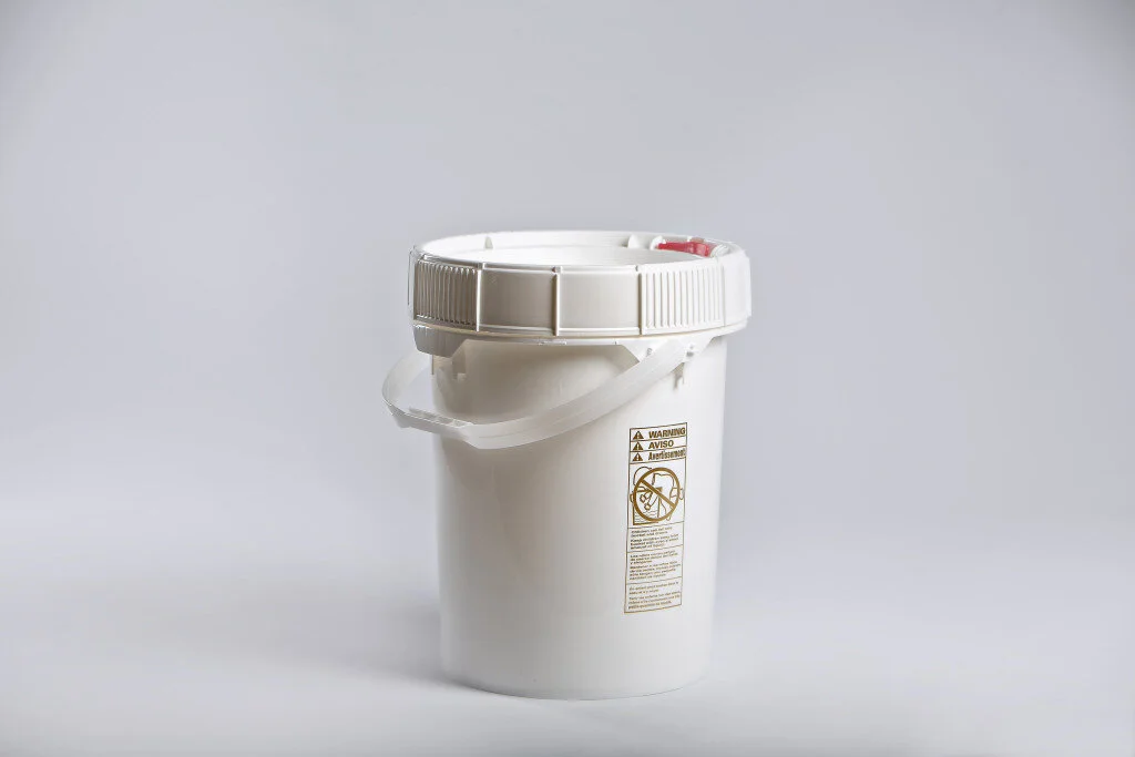 White plastic container with a lid, safety warning label, and handle, used for storing or disposing of hazardous materials.