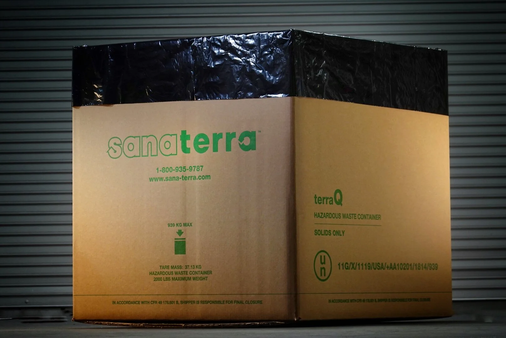 A cardboard hazardous waste container with green text that reads 'sana terra' and 'terraQ', placed against a metal roll-up door background.