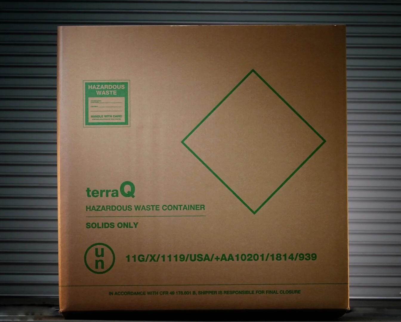 A cardboard hazardous waste container with green text and symbols, marked for solids only, featuring a diamond-shaped symbol and handling instructions.