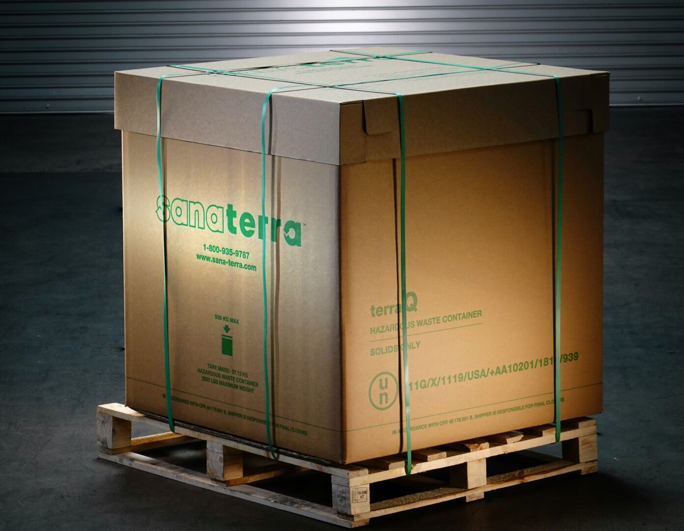 A large cardboard box labeled 'Sanaterra' on a wooden pallet, with green plastic straps securing it, placed on a concrete floor with a metal roll-up door in the background.