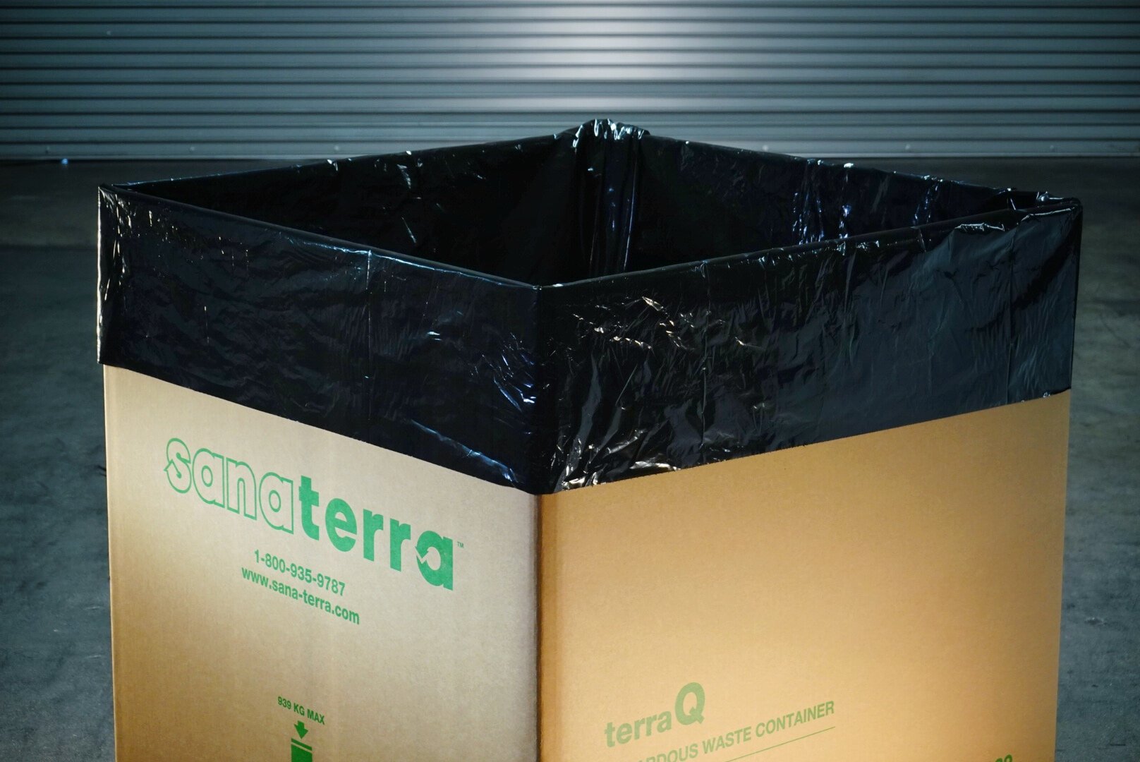 Open recyclable waste container with black plastic liner inside, labeled 'sanaTerra terraQ' in green text, placed on a concrete floor with a gray industrial door in the background.