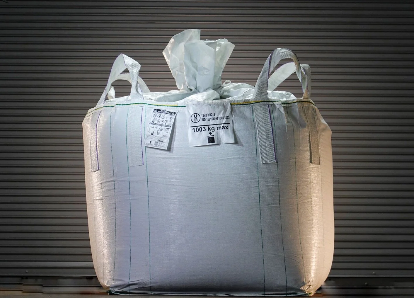 Large white industrial bag with handles, placed in front of a metal shutter, containing a crumpled plastic sheet or bag sticking out from the top.