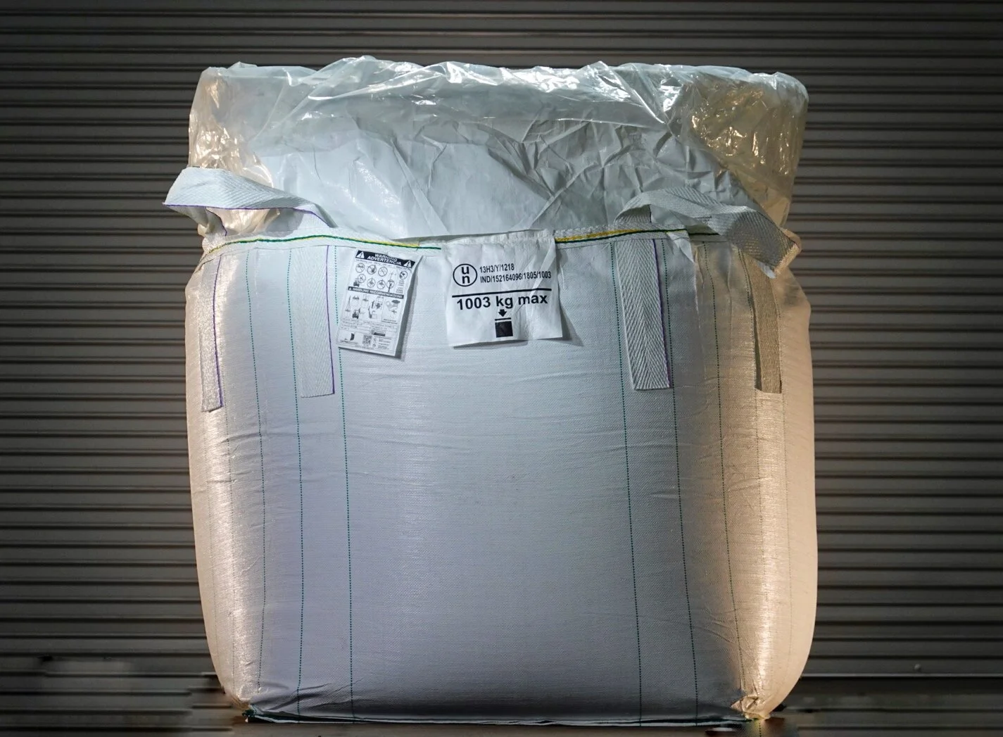 A large white industrial sack with a maximum weight capacity of 1003 kilograms, placed in front of a gray metallic rolling shutter.
