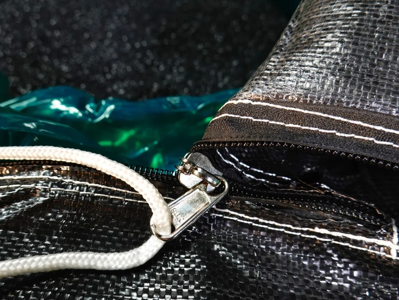 Close-up of a zipper on a black bag with textured, shiny material, with white and black cords, and reflective green and blue plastic in the background.