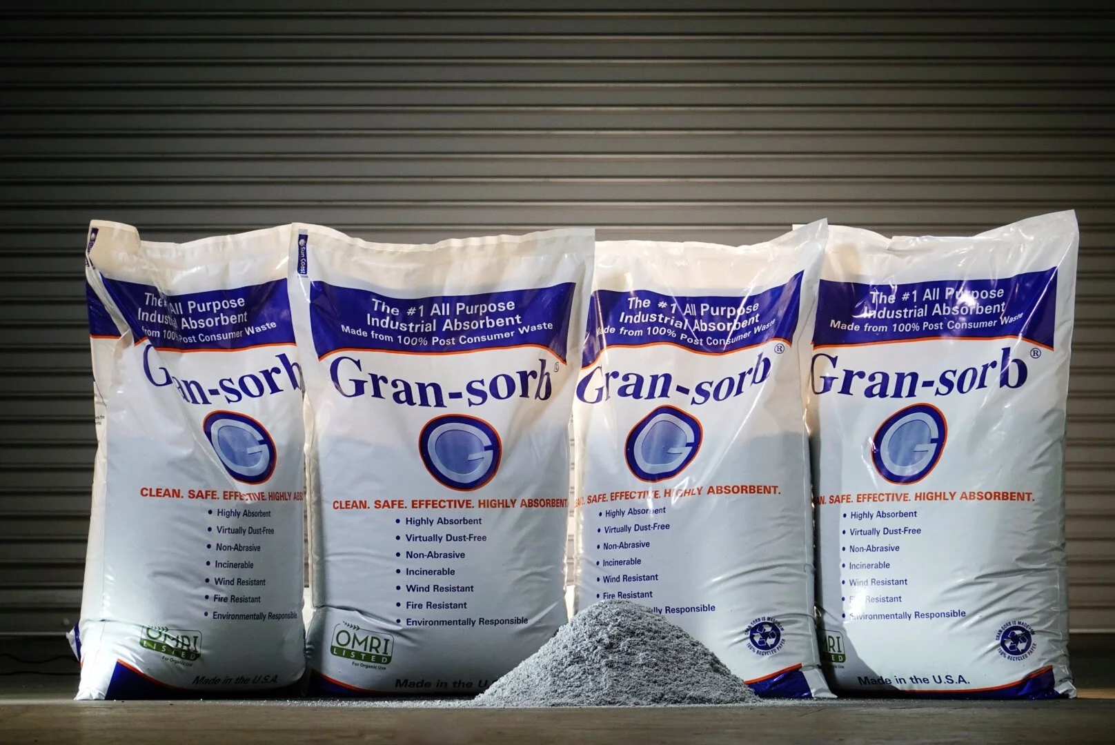 Four bags of Gran-sorb industrial absorbent powder with a small pile of the gray powder in front of them, set against a metallic background.