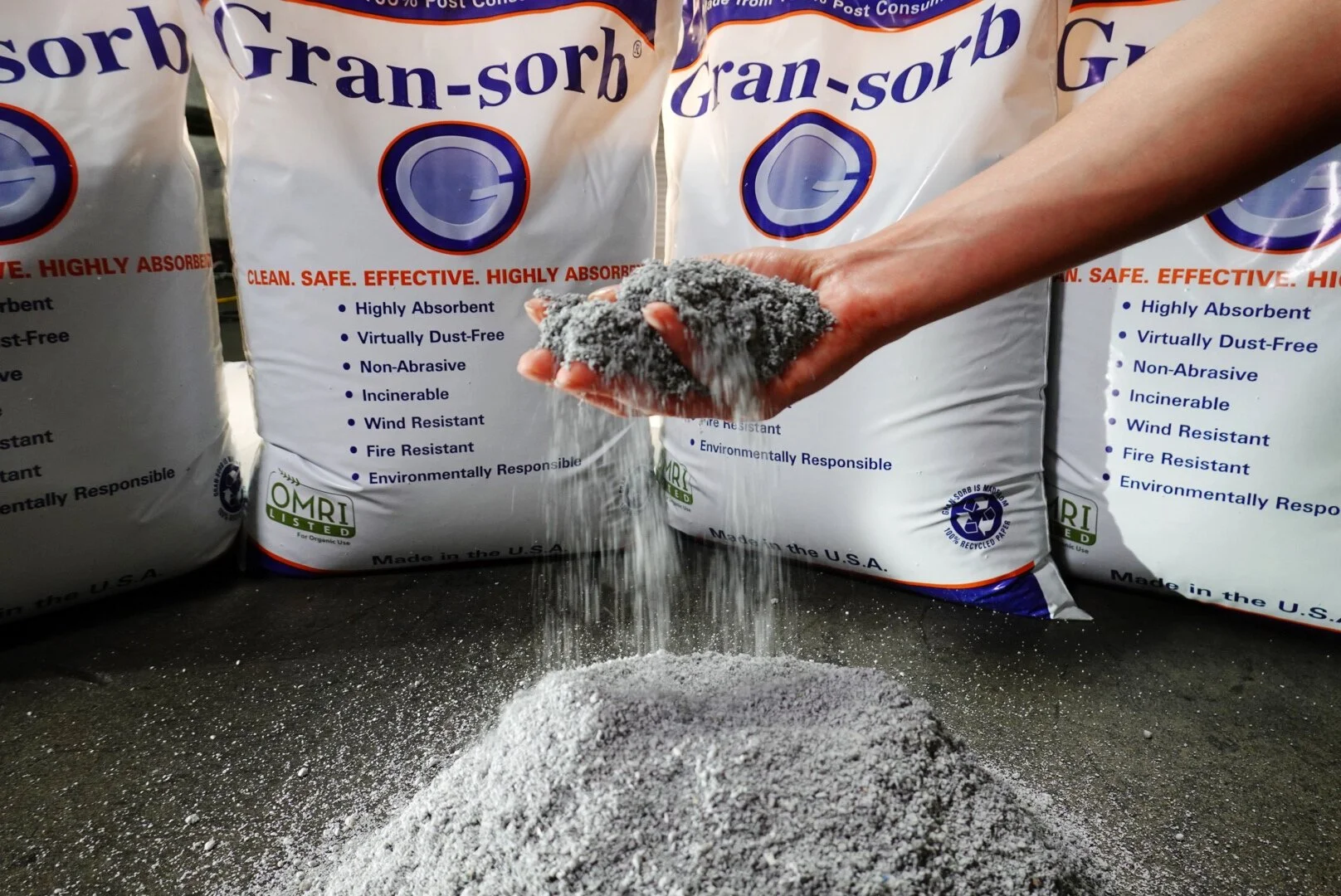 Person holding gray granulated absorbent powder, with bags of gran-sorb in the background.