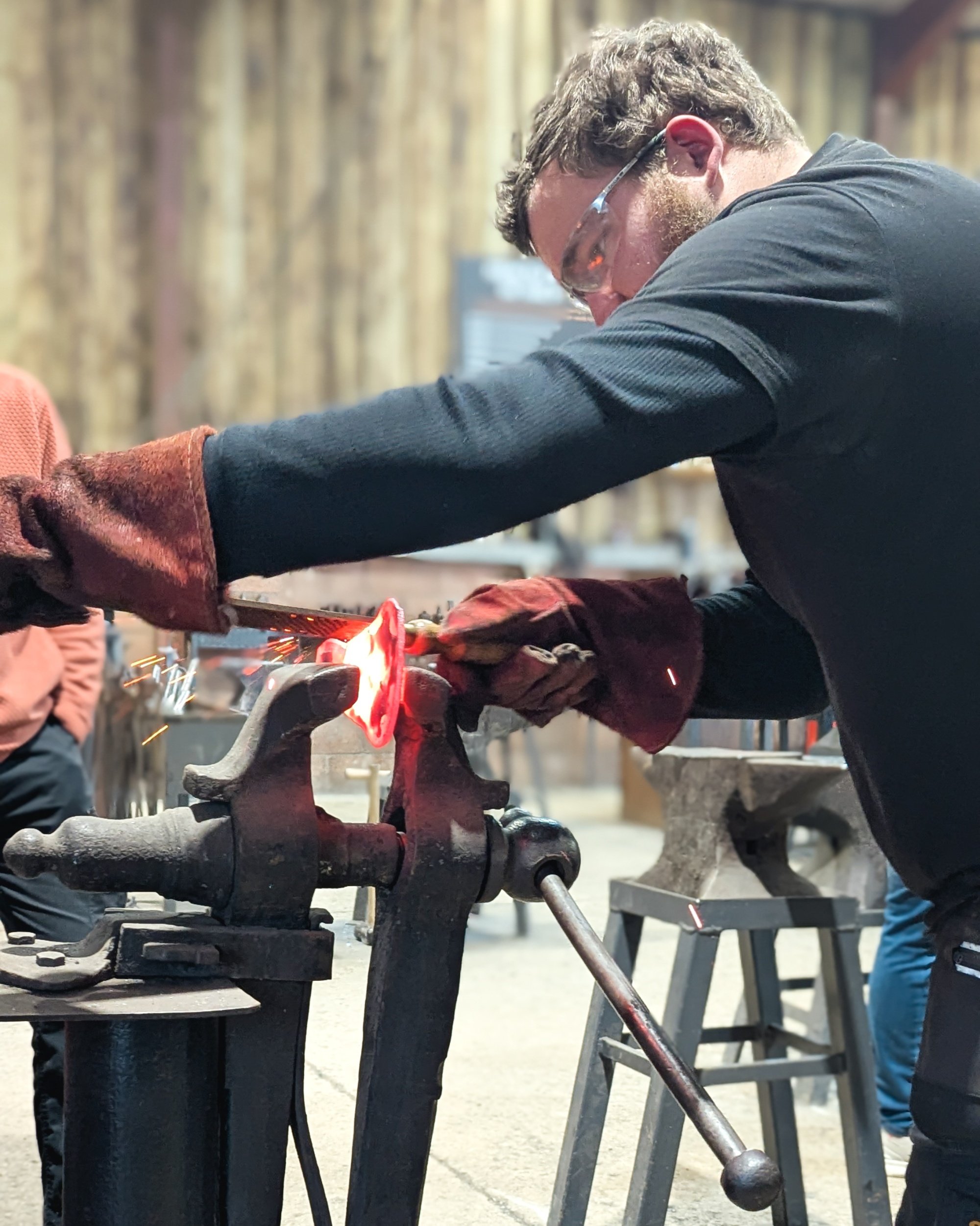 Close-up of hot rasping in progress; a metal rasp removes material from a hot steel axe head in a vice to refine its curved profile before it cools.