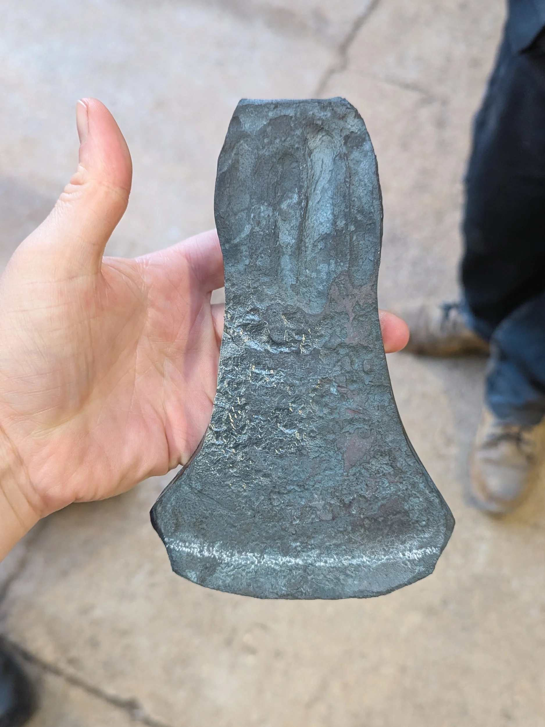 A close-up of a student’s hand holding a cooled, forged steel axe head at Oldfield Forge, showing the refined profile and tapered edge achieved after the initial shaping stages.