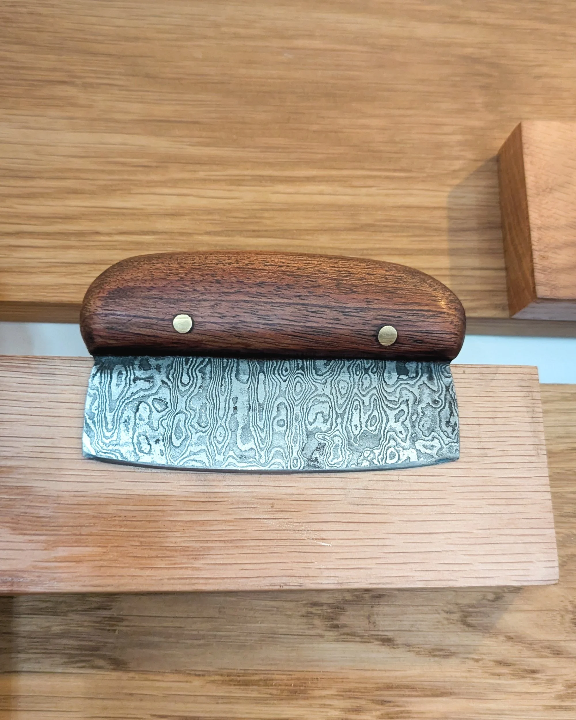 A luxury pizza cutter featuring a wavy Damascus steel blade and a handcrafted wooden handle secured with decorative gold pins, showcasing artisanal kitchenware at Oldfield Forge.