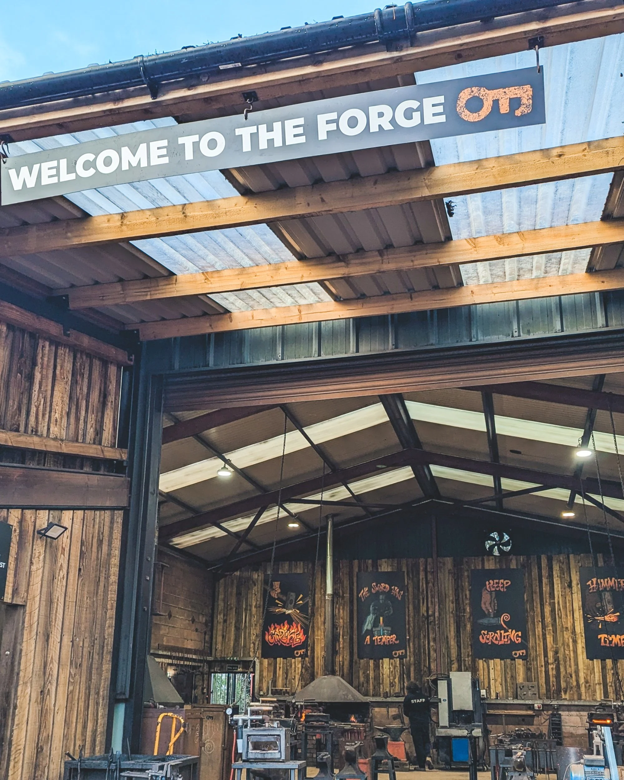 The entrance to the Oldfield Forge open-barn workshop, showing a large branded sign, traditional coal forges, anvils, and blacksmithing tools ready for a class.