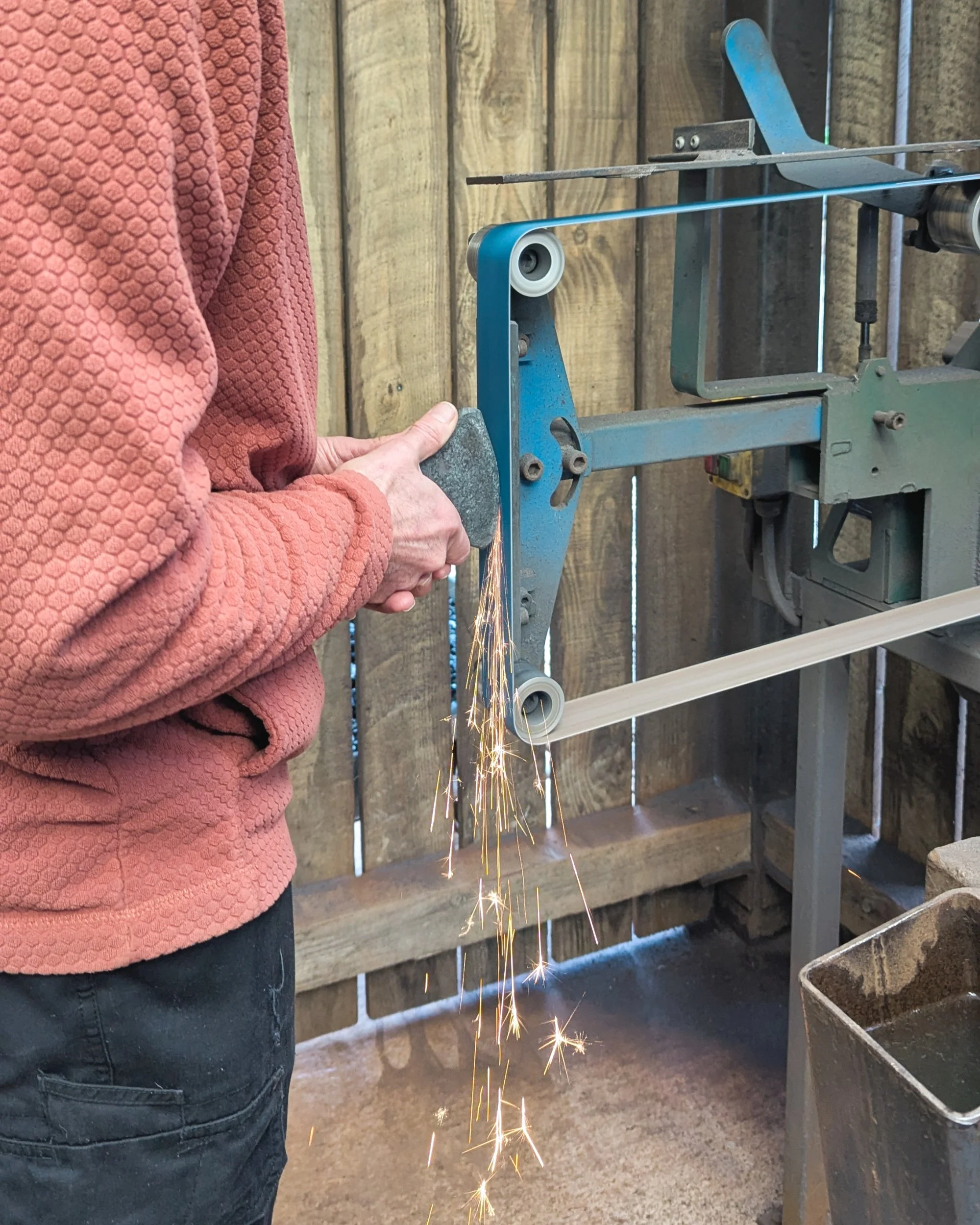 A dynamic action shot of a blacksmith using a high-speed belt sander to refine the front edge of an axe head, with a vibrant shower of sparks flying as the rough forge scale is ground away.