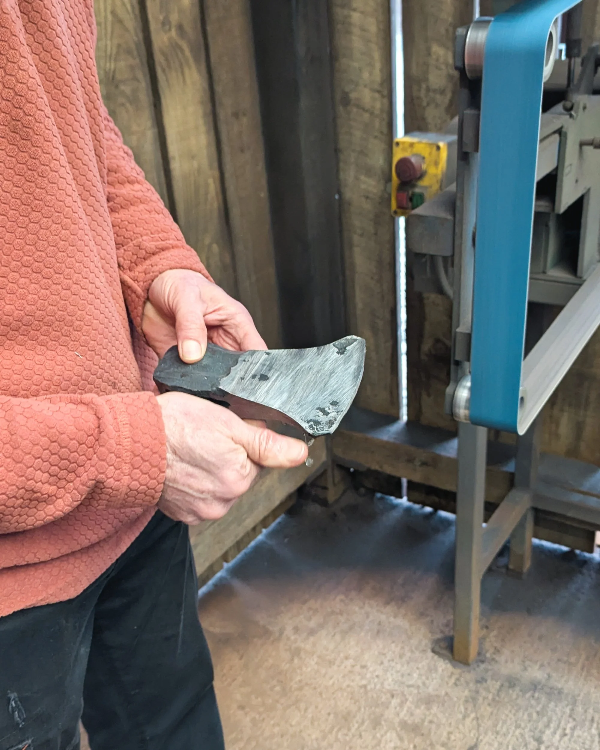 A blacksmith standing next to a belt sander, carefully inspecting the polished flat face of a steel axe head. The bright, reflective surface reveals the smooth finish and the absence of marks or dimples.