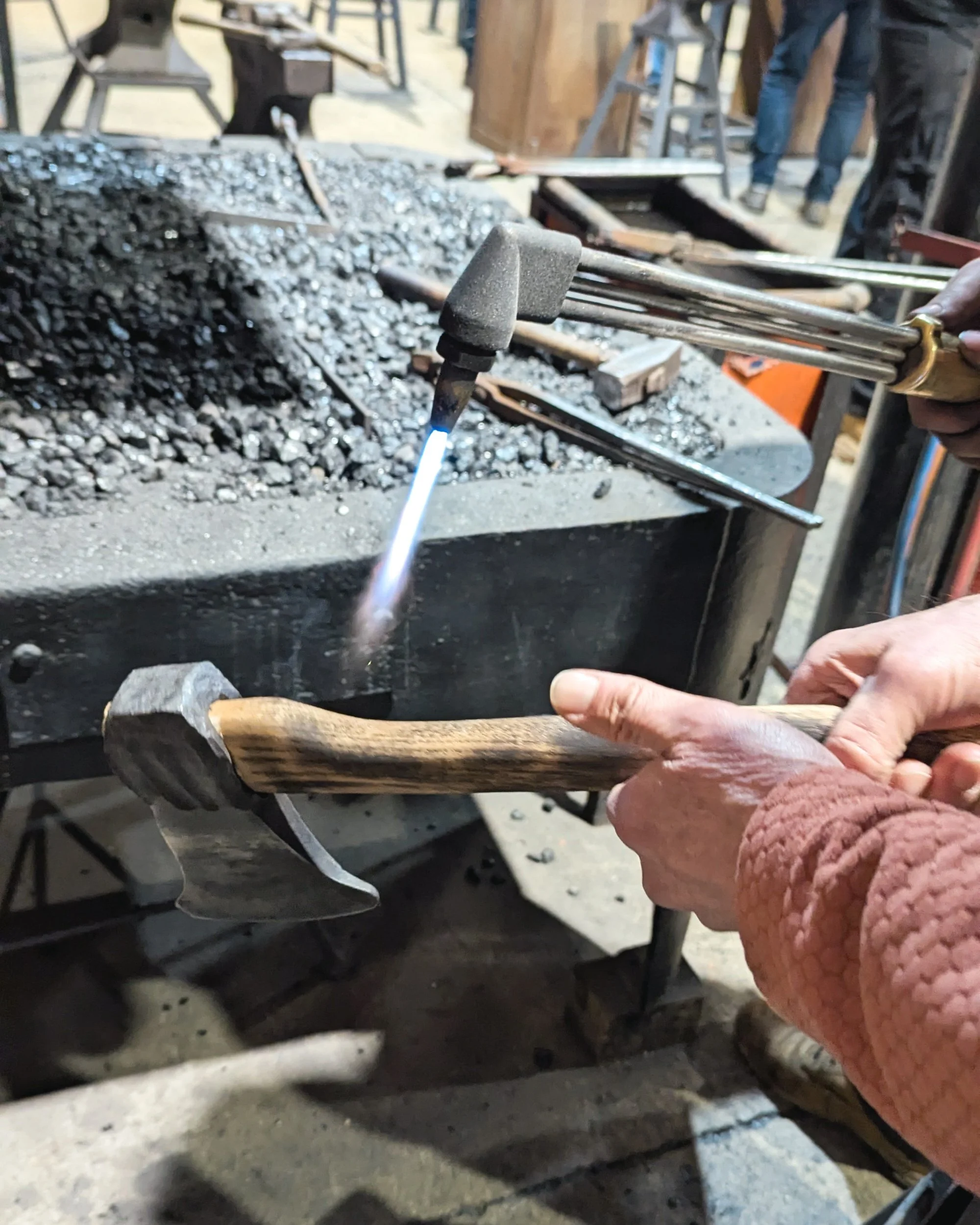 A blacksmith using a gas blow torch to lightly char a wooden axe handle, bringing out the natural grain patterns and creating a rustic, burnt finish.