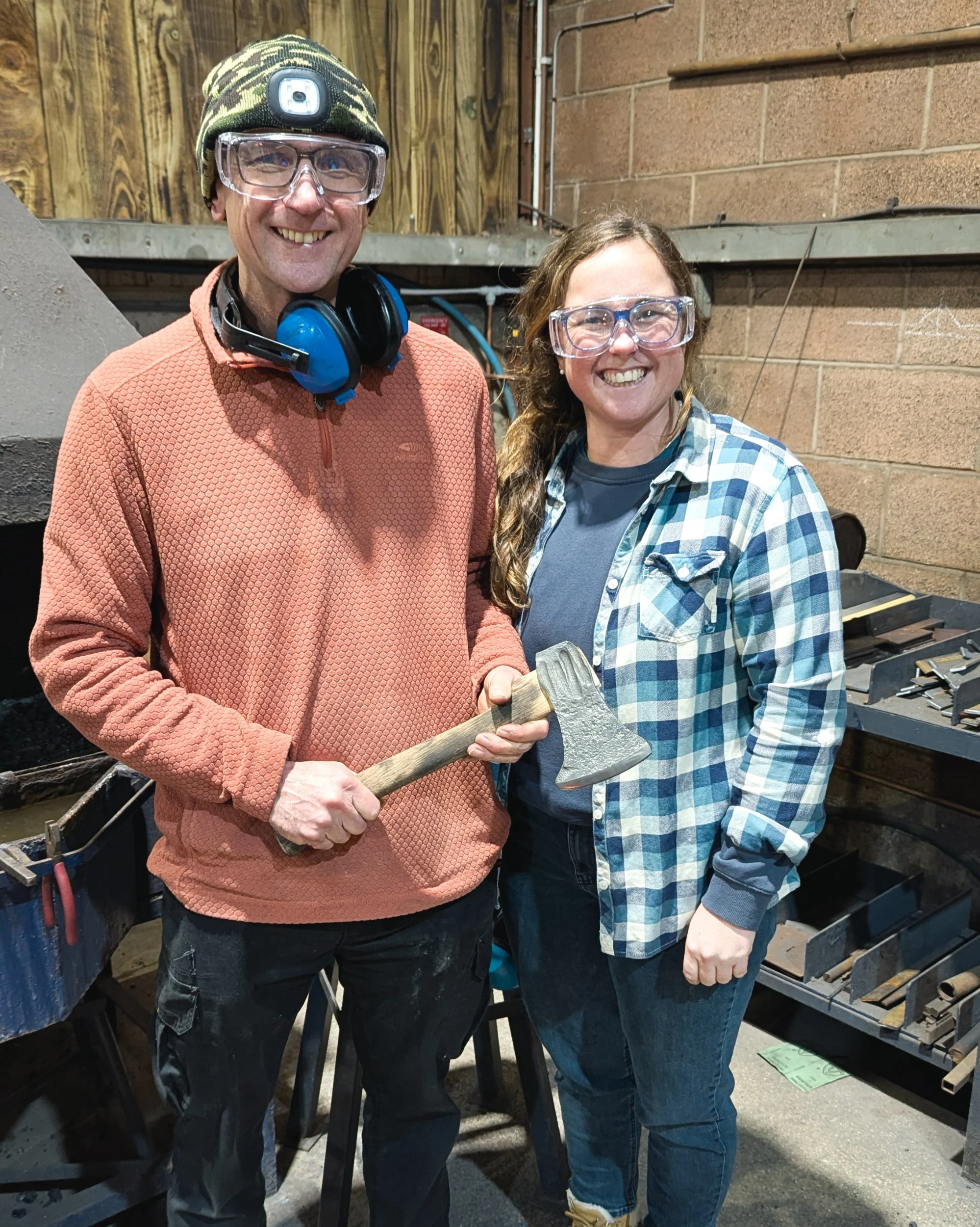 A father and daughter smiling together in the workshop, proudly holding the hand-forged axe they created during their blacksmithing course at Oldfield Forge.