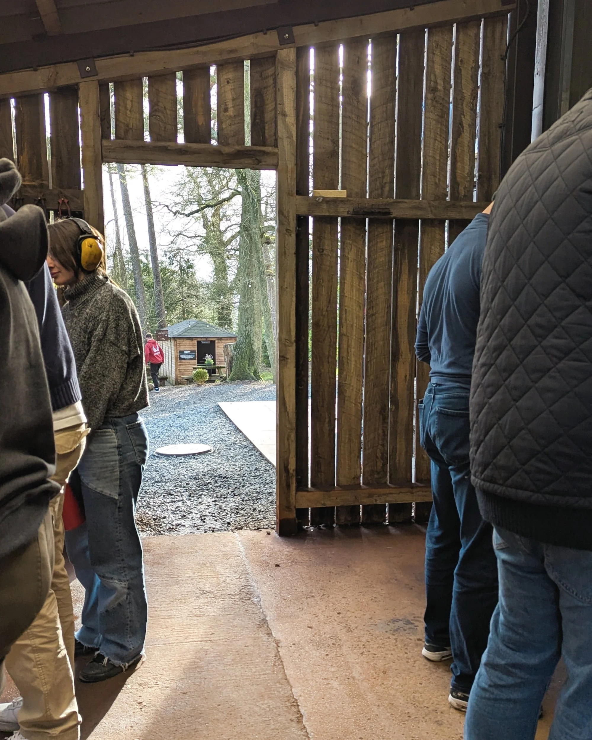 An interior view of students working at belt sanders inside the small timber-framed workshop barn, with large open doors offering a scenic view out to the green grounds and woodland.