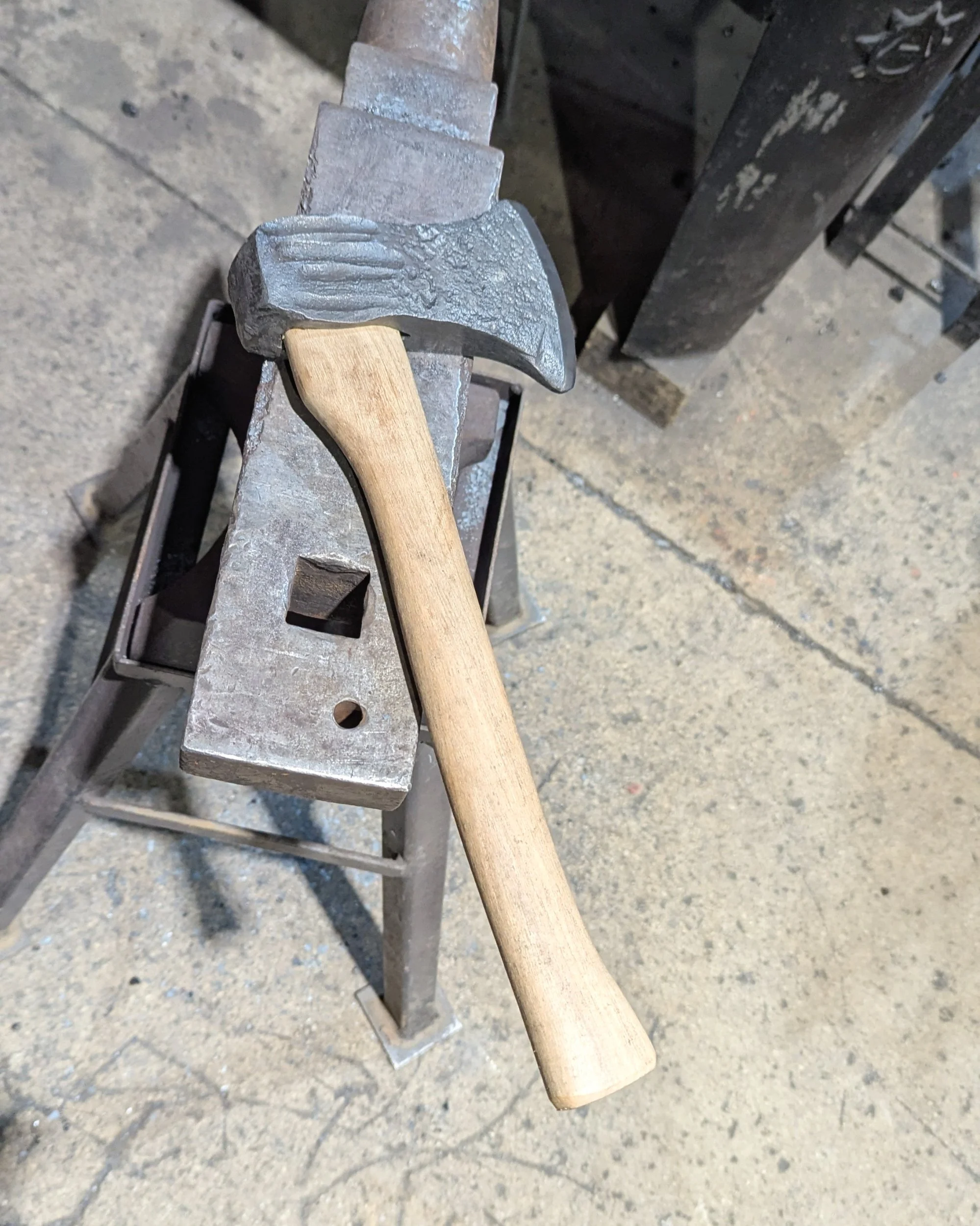 A fully assembled, hand-forged axe with a polished Damascus edge resting on a blacksmith's anvil at Oldfield Forge, symbolizing the completion of the crafting process.