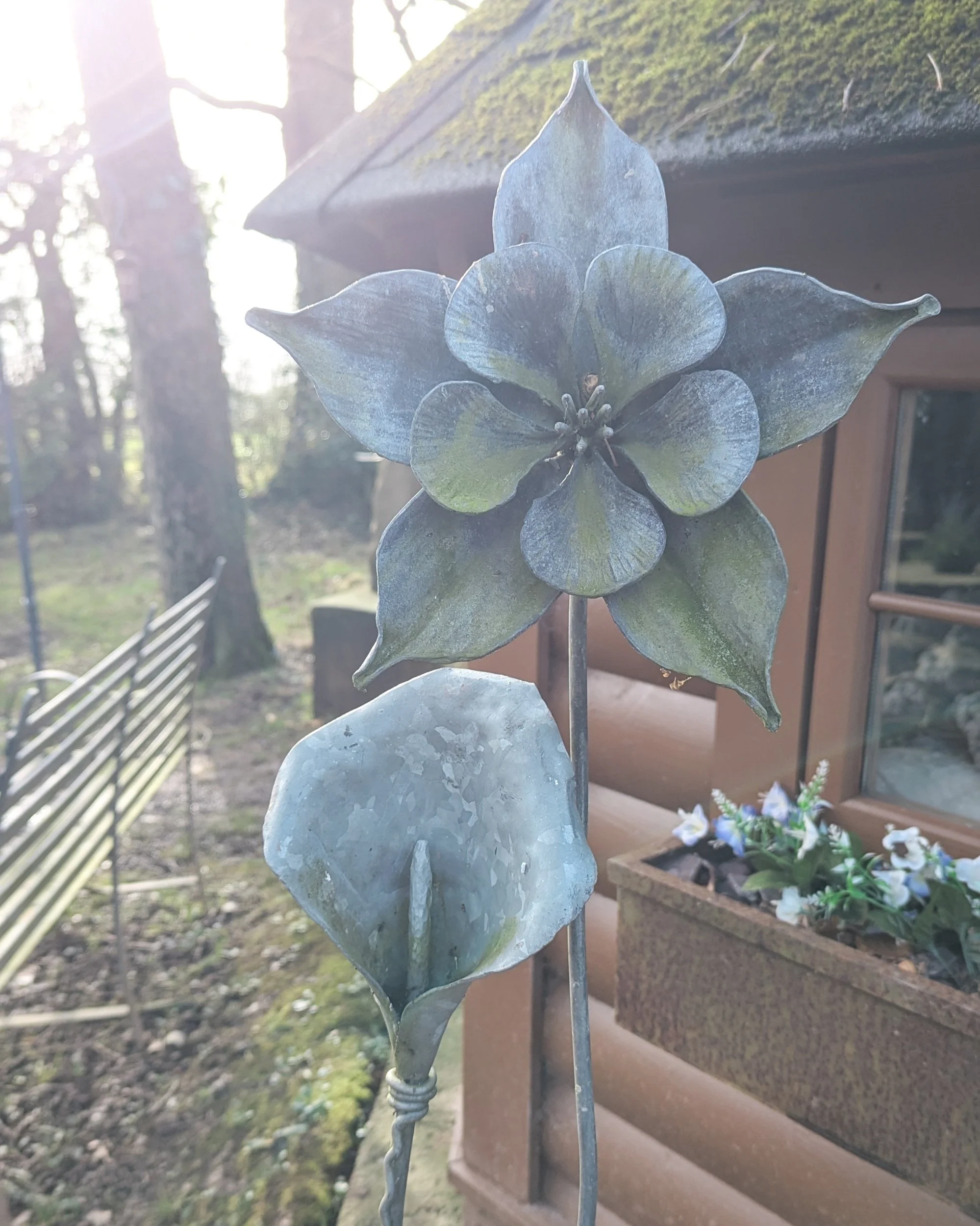 A macro close-up of an intricate, hand-forged metal flower sculpture at Oldfield Forge, showing the textured petals and hammered details of the steel artwork in a garden setting.