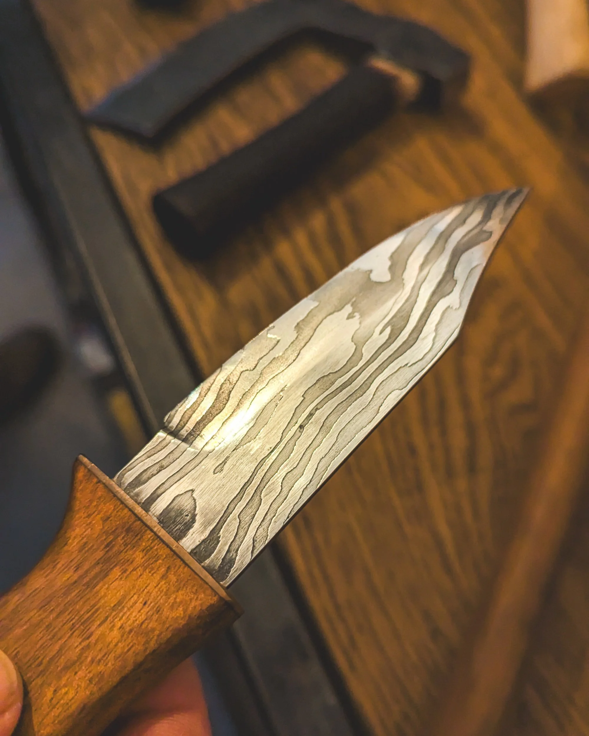 Extreme close-up of a hand-forged Damascus steel knife blade at Oldfield Forge, showing the intricate, wavy wood-grain patterns of the layered metal.