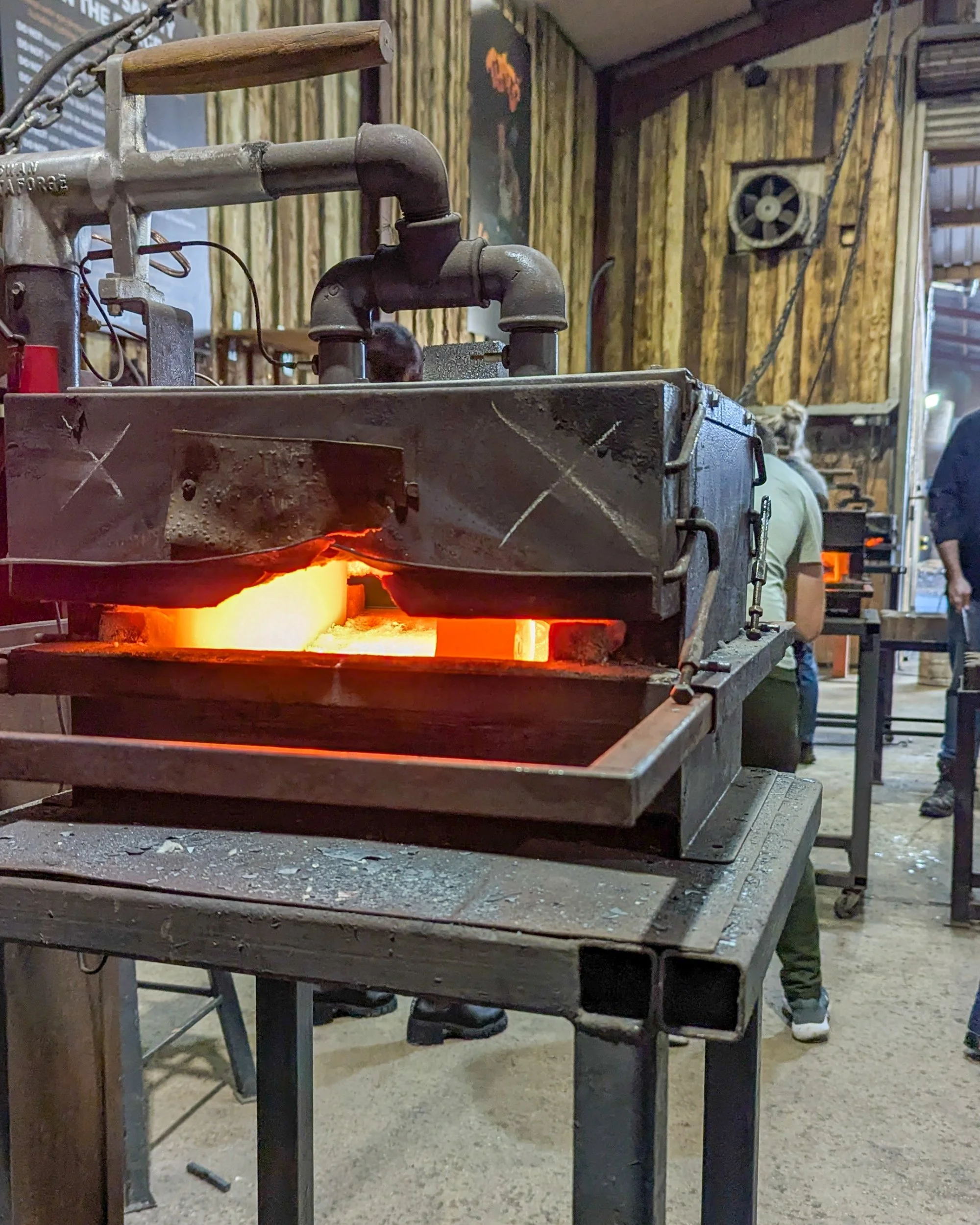 A close-up view inside a gas forge oven showing a square billet of steel glowing vibrant orange-yellow, heated to over 600°C and ready for the anvil.