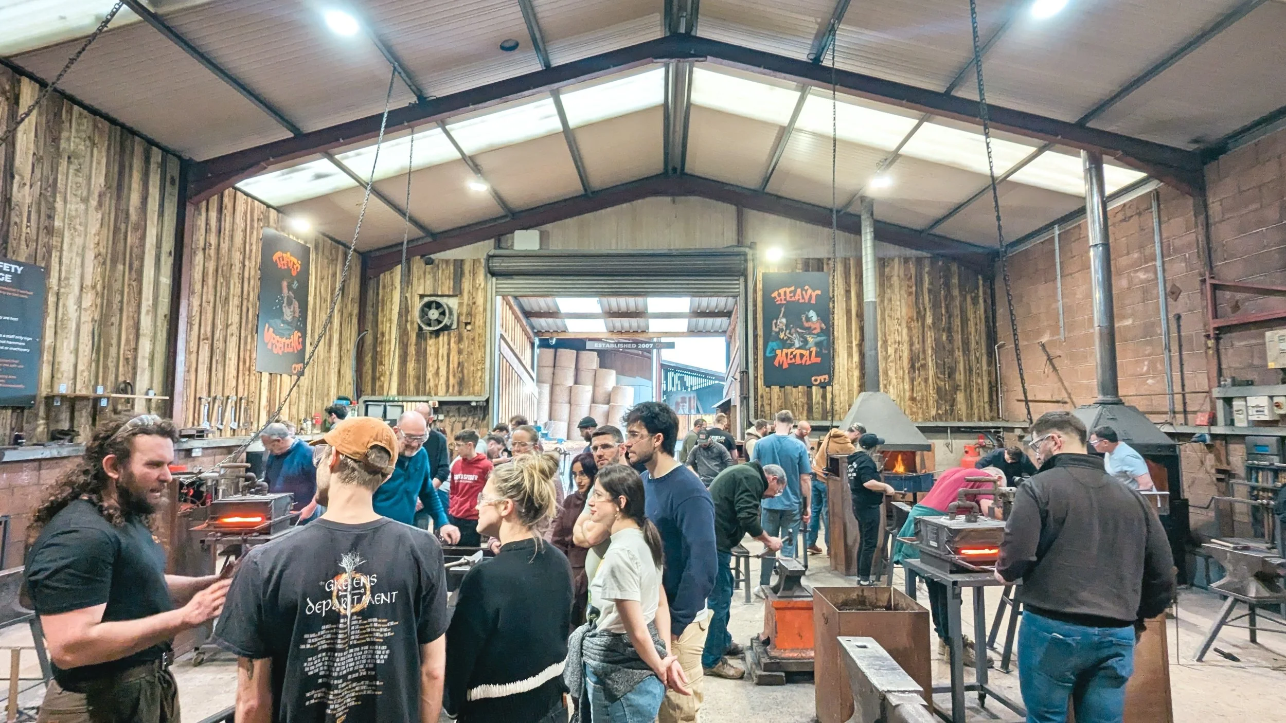 A busy, high-energy workshop at Oldfield Forge with groups of people crafting various items, including hand-forged swords, daggers, and kitchen knives during an introductory blacksmithing course.