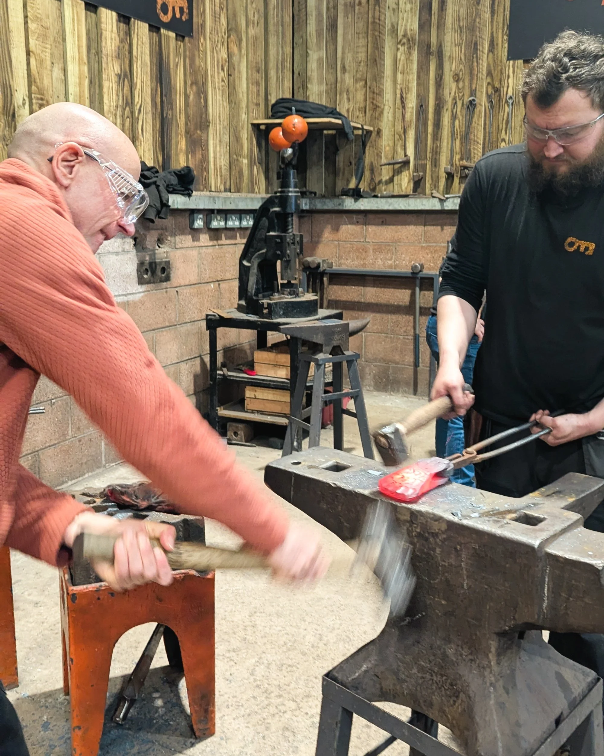 An energetic, motion-blurred action shot at Oldfield Forge capturing the powerful, fast-paced rhythm of a blacksmith’s hammer striking a tool.