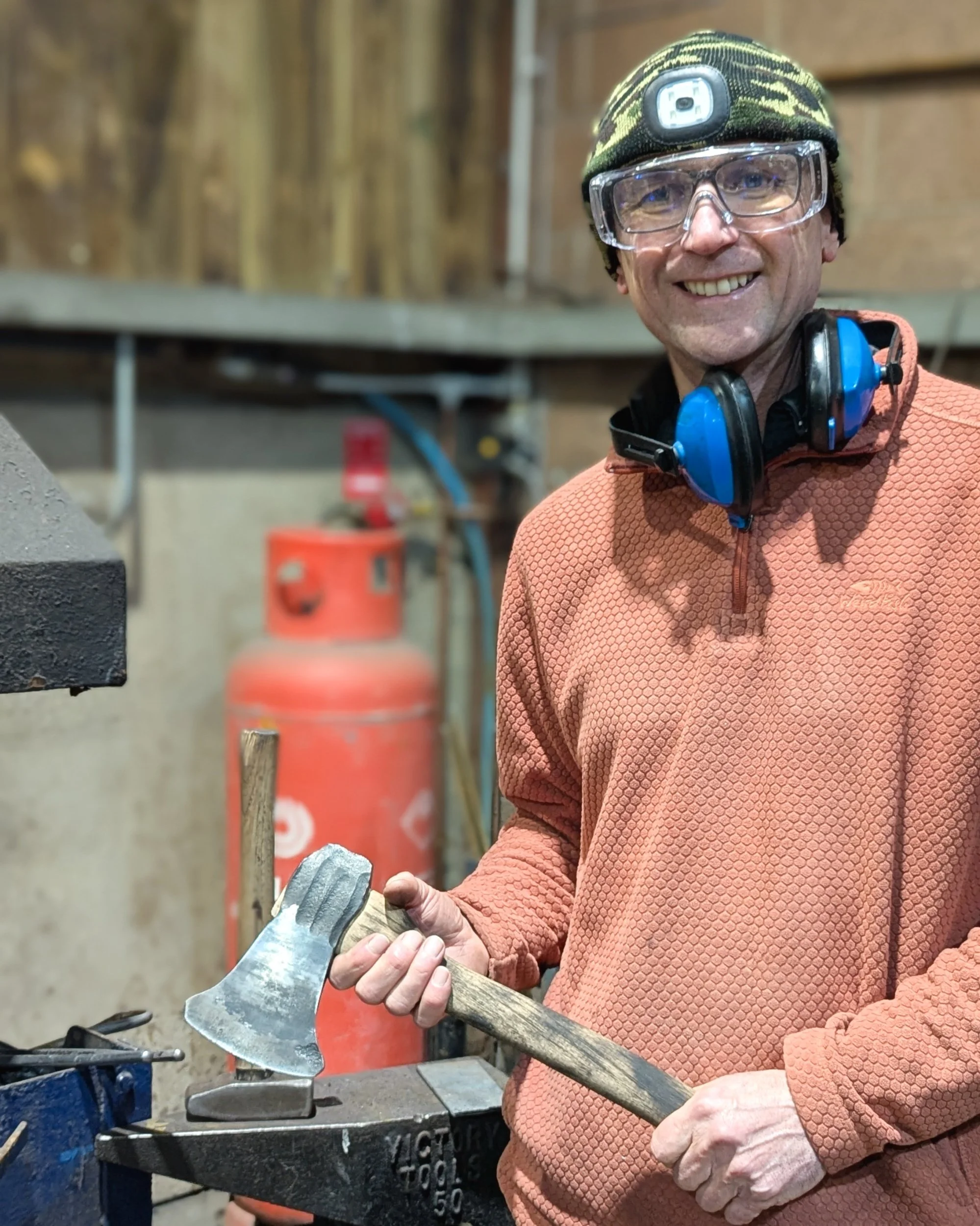 A student standing in the Oldfield Forge workshop, smiling and holding their completed hand-forged axe, showcasing the charred handle and polished Damascus steel head.