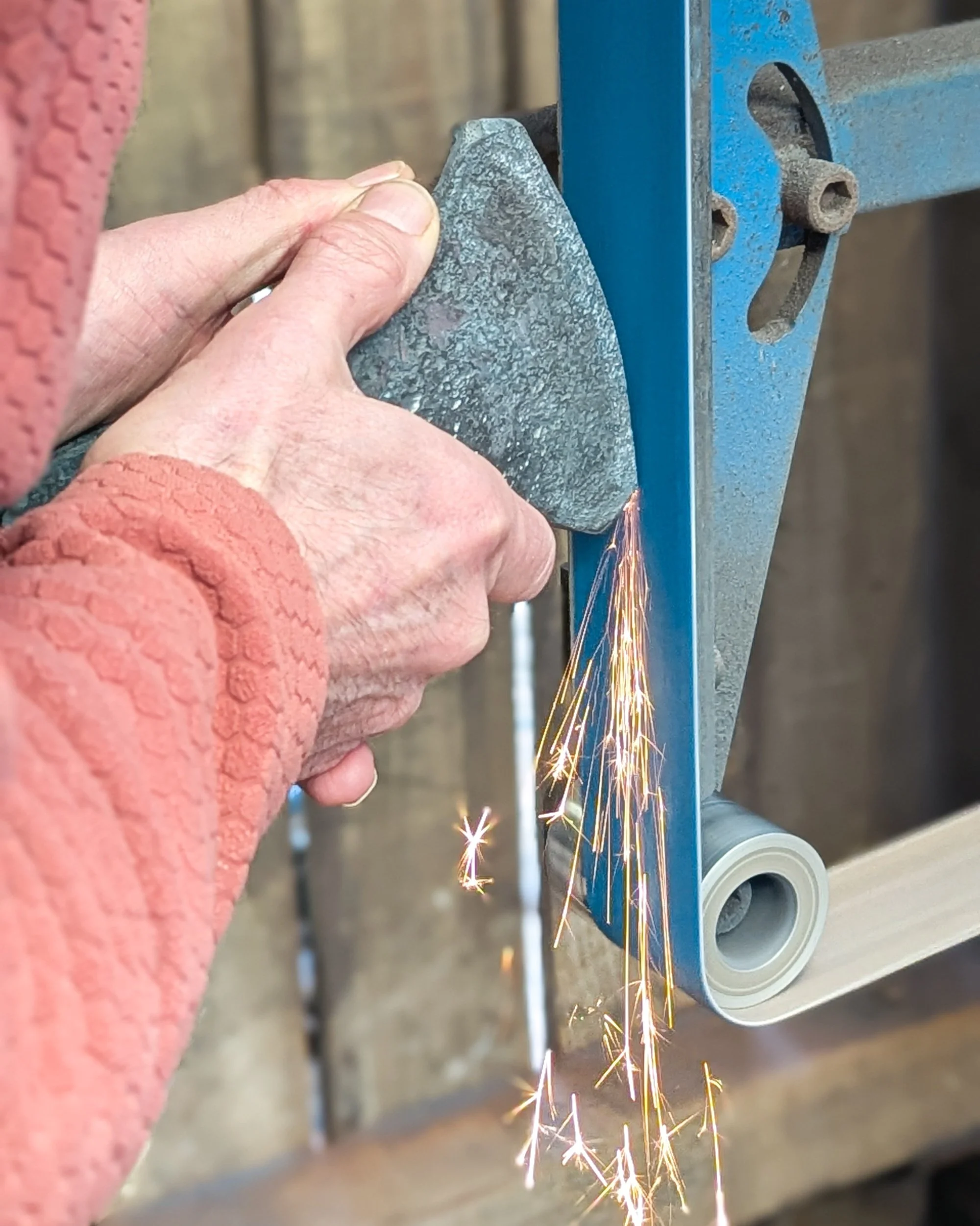 A close-up showing the precise shaping of an axe's primary bevel on a belt sander, capturing the friction and sparks created as the metal is ground into a sharp, clean profile.