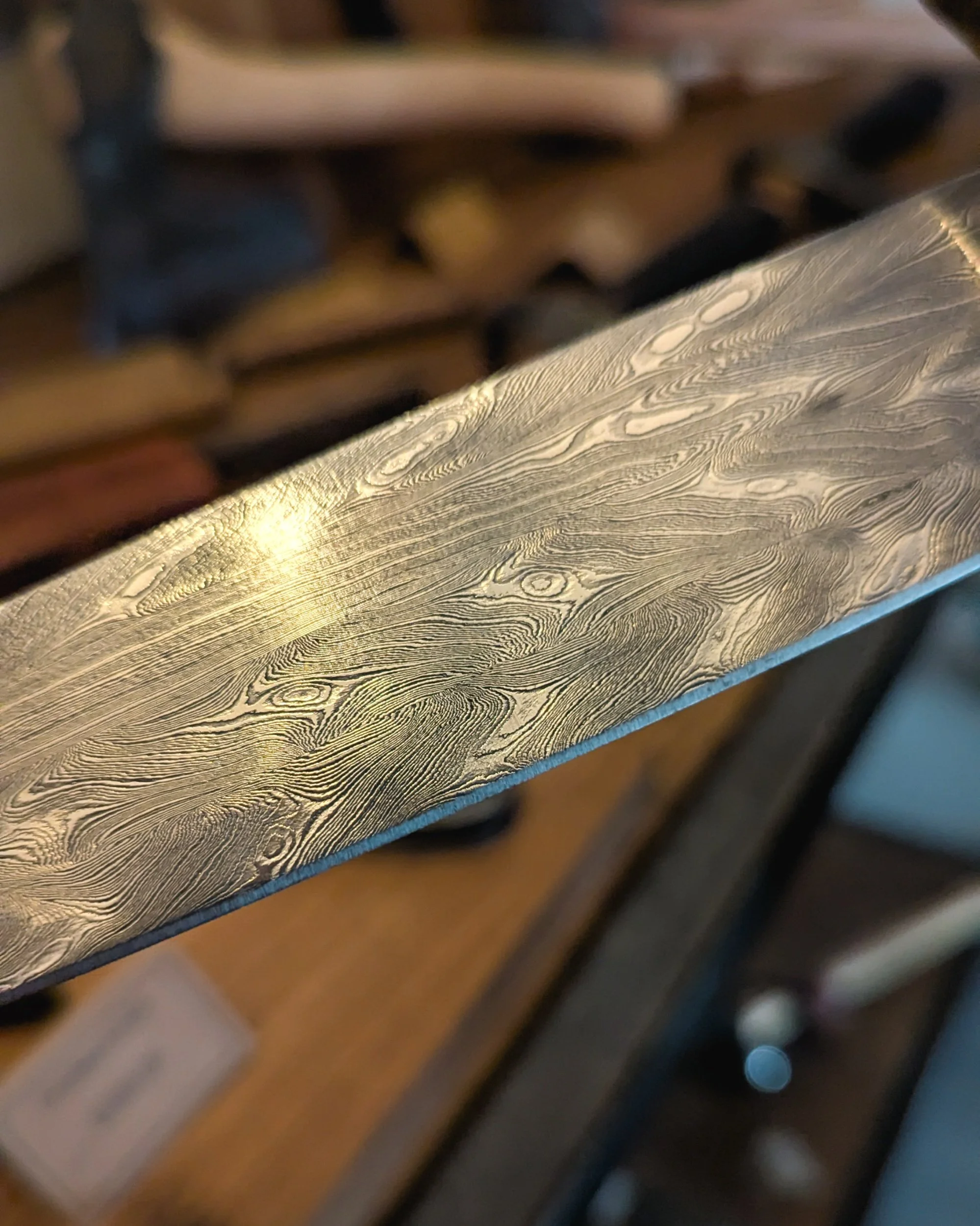 A stunning close-up of a custom-forged knife blade featuring an intricate feather Damascus pattern, where the steel layers mimic the organic symmetry of a bird's feather.