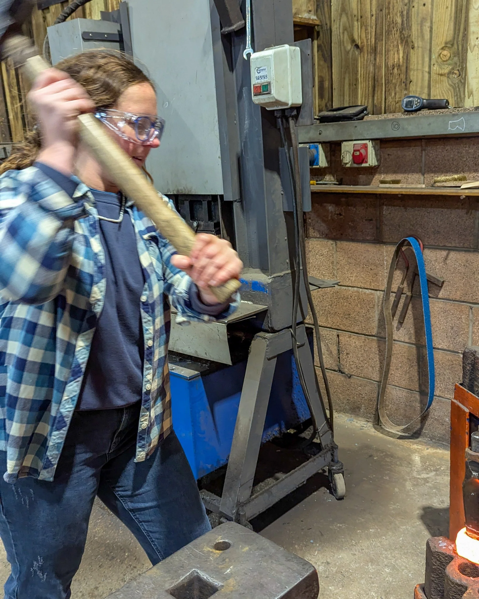 A dynamic action shot of a blacksmith swinging a heavy hammer to strike a metal drift, driving it through a glowing orange axe head on an anvil.