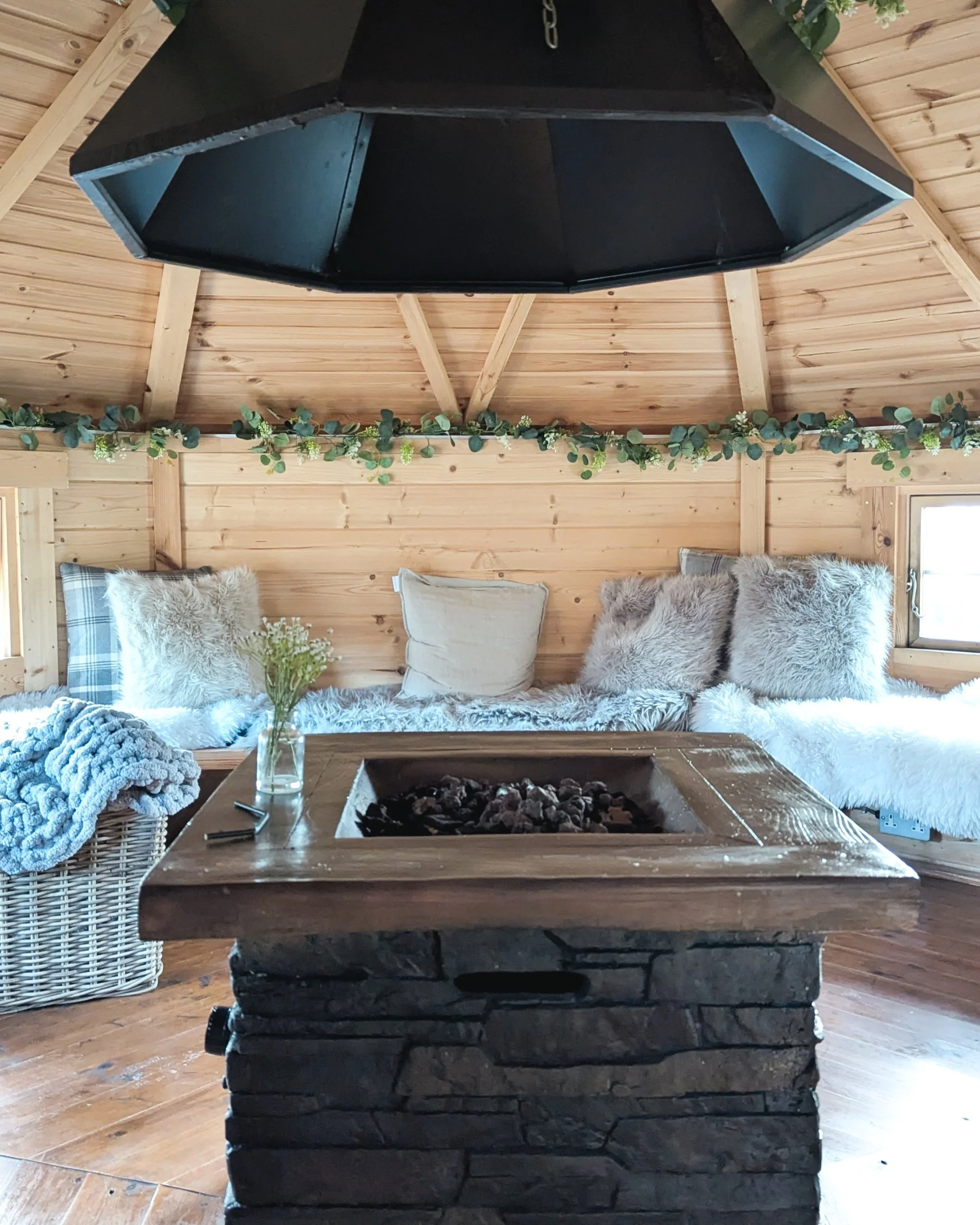 The interior of a modern wooden lodge at Oldfield Forge featuring a central fire pit, angular architectural walls, and cozy benches styled with plush cushions and warm throws.