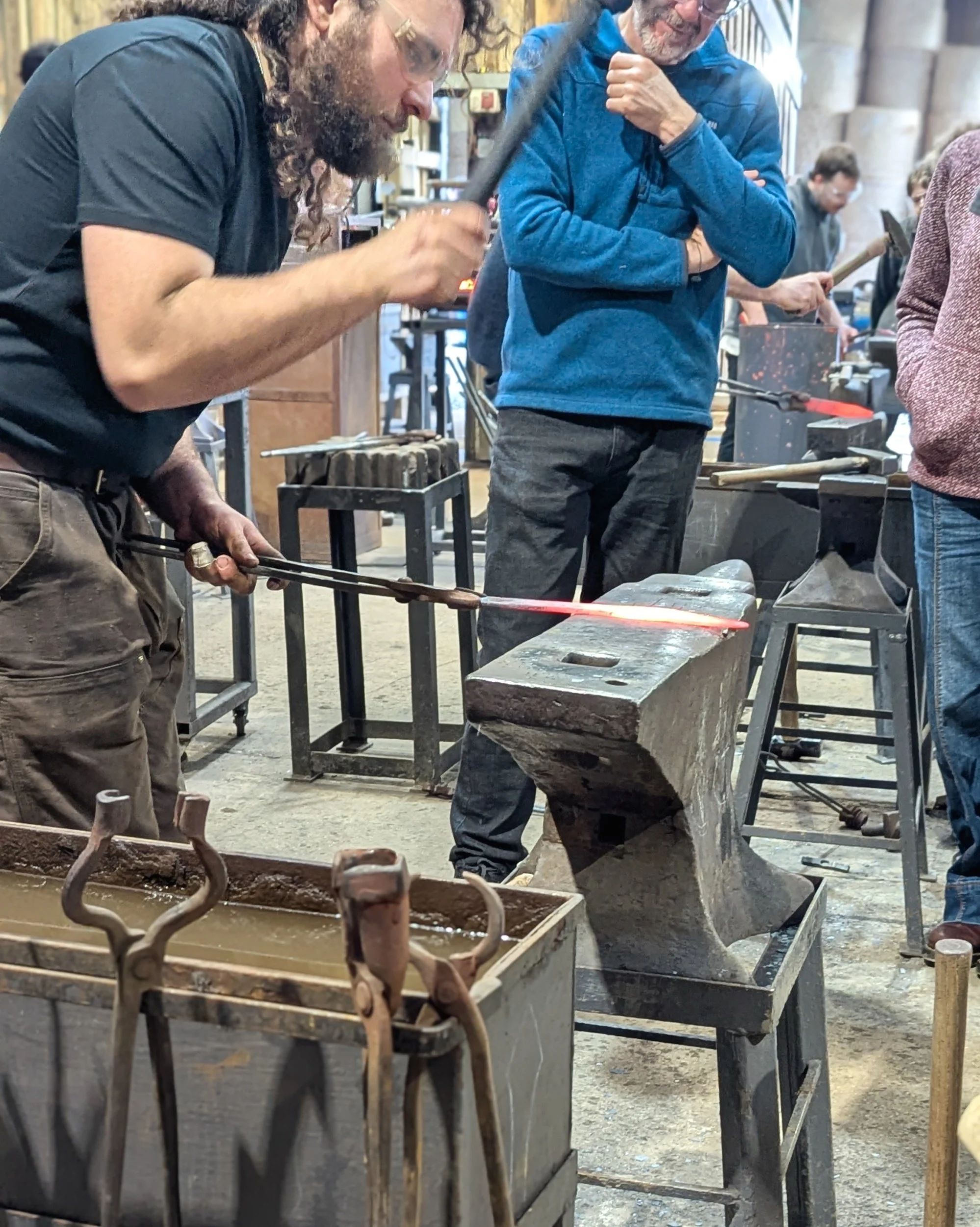 A professional blacksmith instructor at Oldfield Forge demonstrates the technique for flattening a red-hot dagger blade with a hammer while a group of students watches closely.