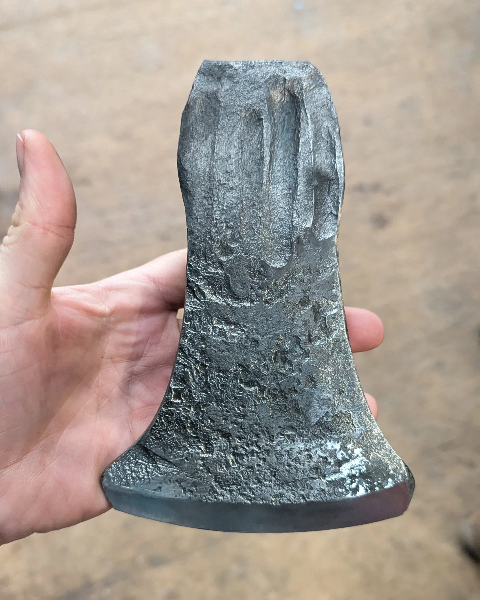 A hand holding a recently forged axe head that has been wire-brushed to a clean grey finish, featuring a bright, shiny ground front edge that highlights the blade's profile.