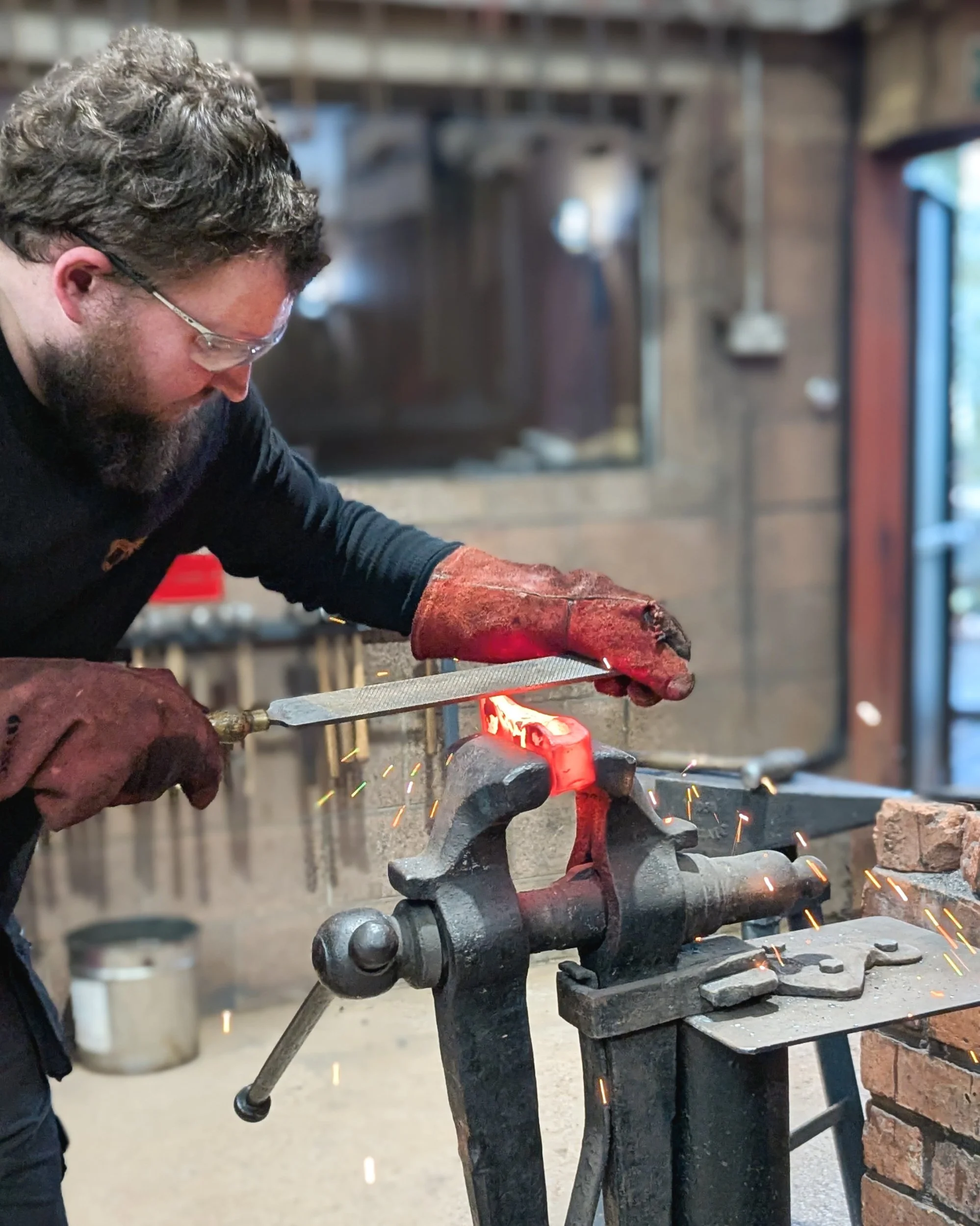 Close-up of hot rasping in progress; a metal rasp removes material from a hot steel axe head in a vice to refine its curved profile before it cools.