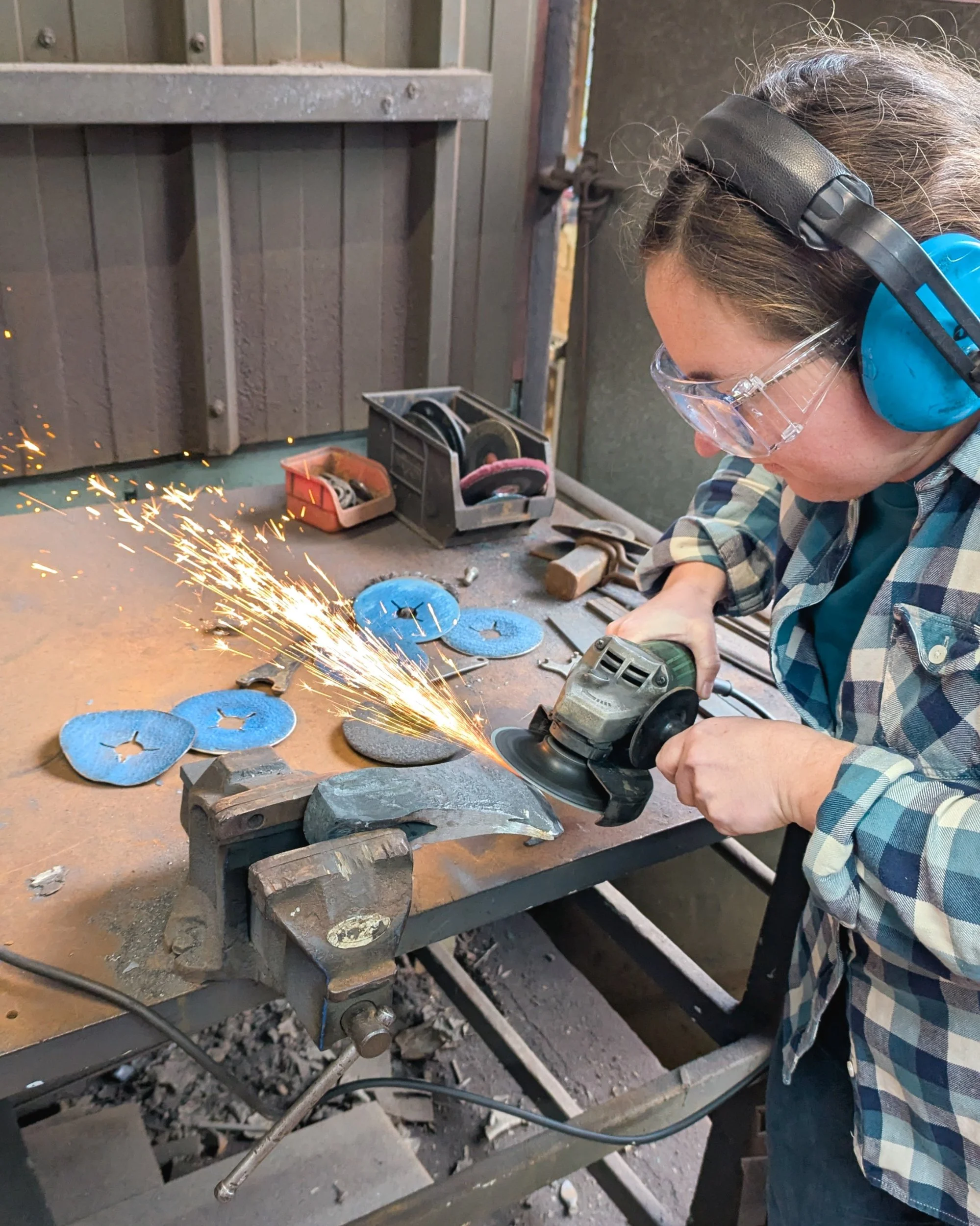 Using a power grinder to add a sharp edge to a forged axe head held in a workshop vice.