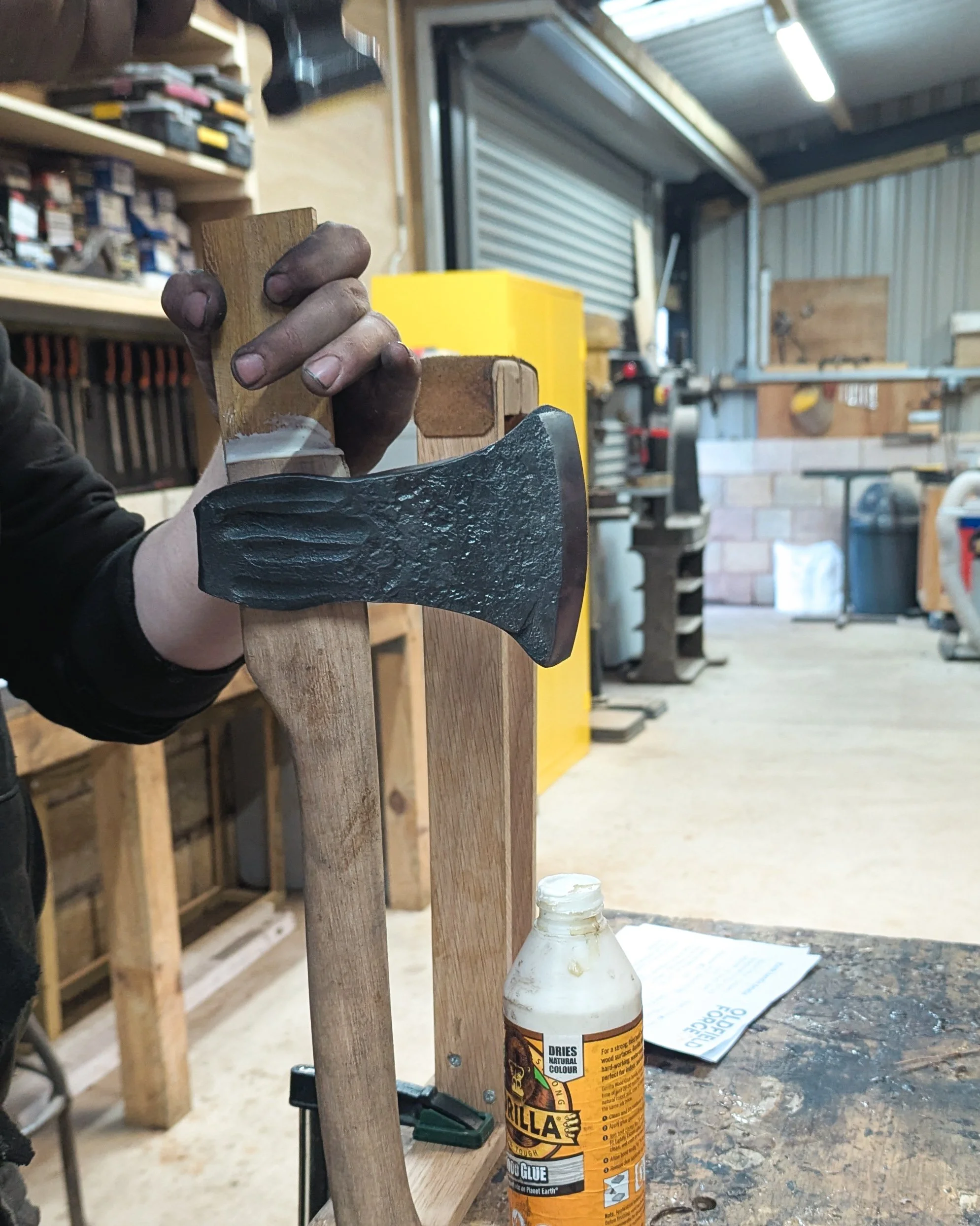 An action shot of a hammer striking a wooden wedge into the top of an axe handle, expanding the wood within the eye of the forged steel head to create a permanent, secure fit.