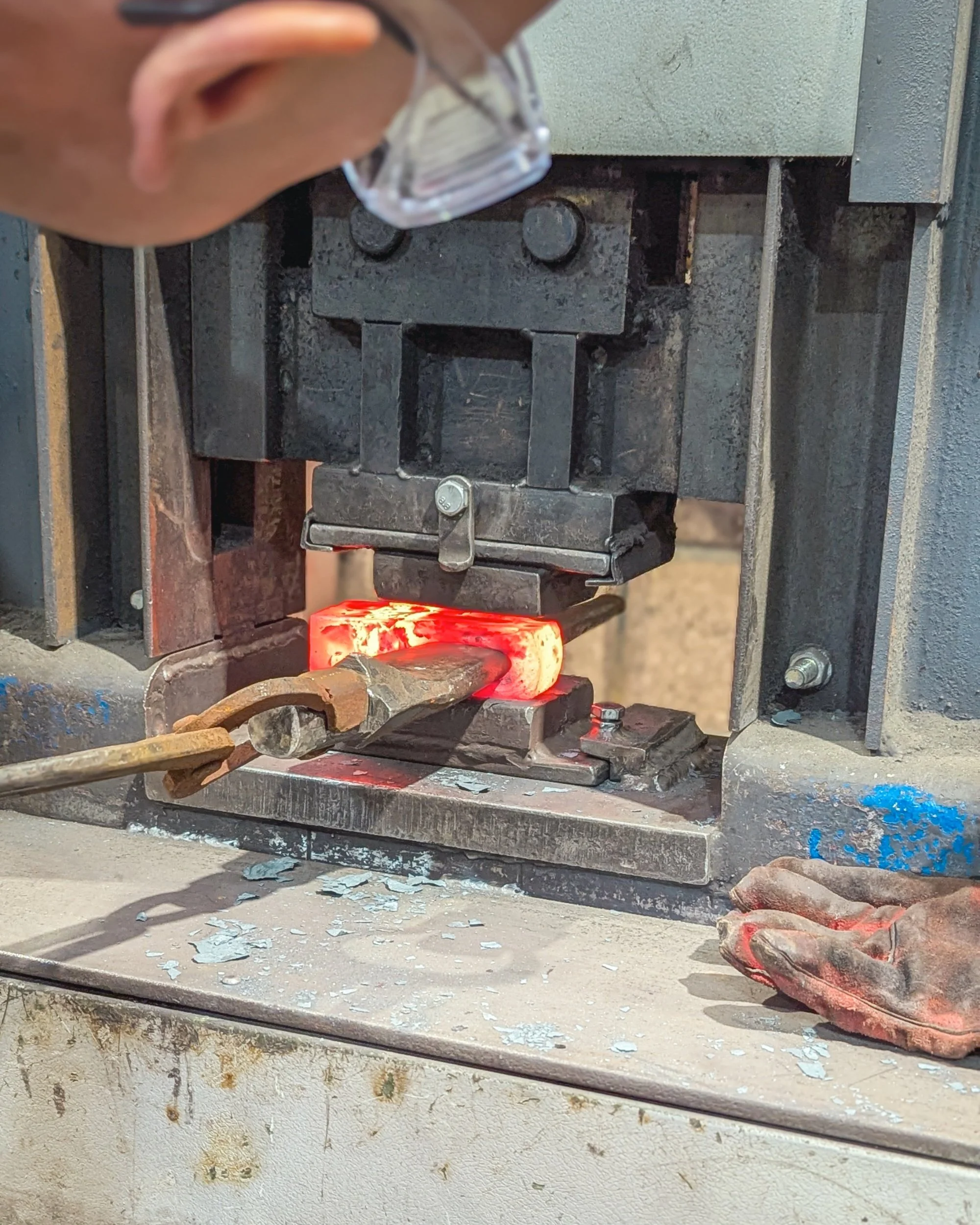 A tight close-up of a hydraulic press die crushing a glowing yellow-hot steel billet, showing sparks and metal scale flaking off under the intense pressure.