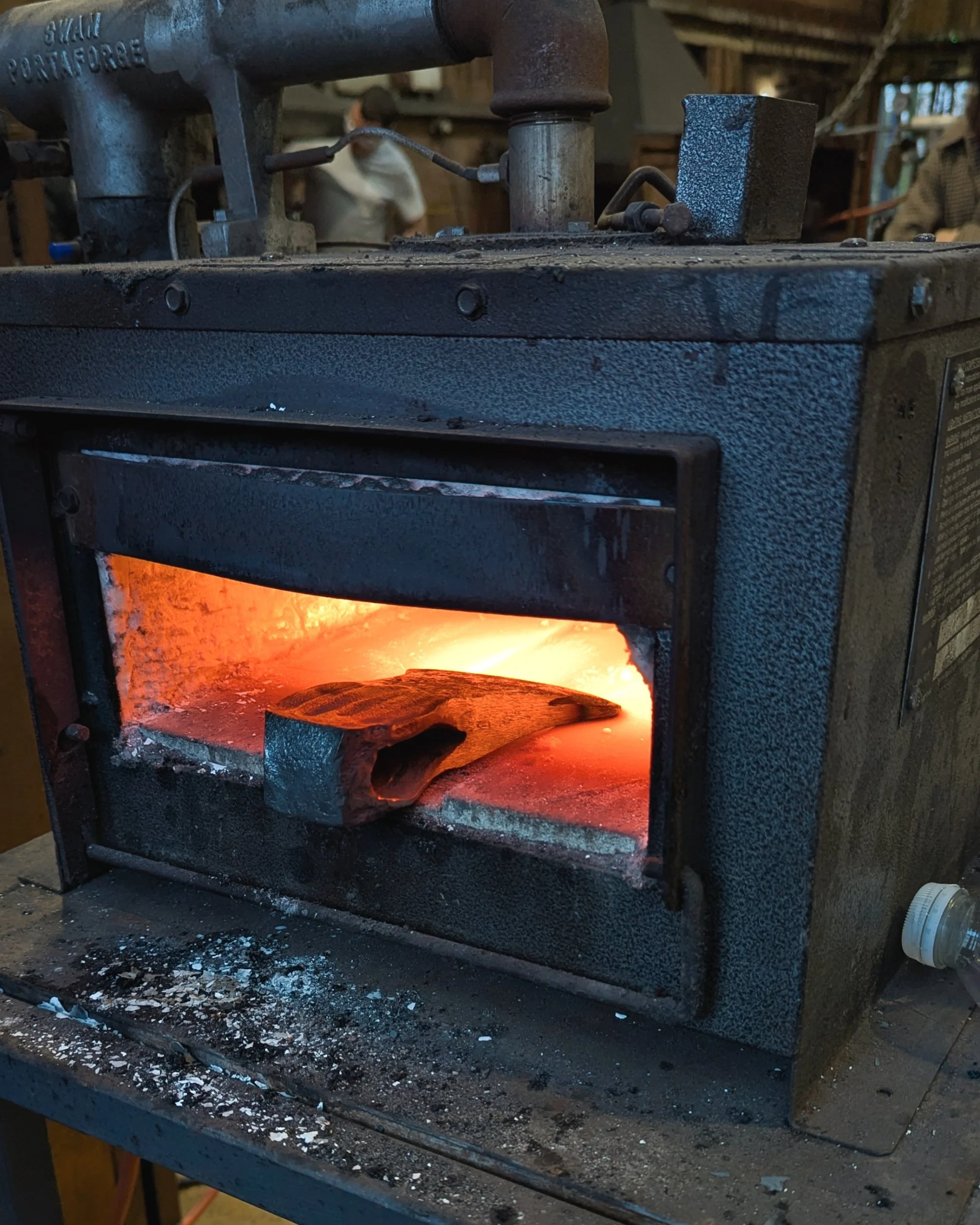 An axe head resting inside a glowing orange gas forge, with the focus on the cutting edge as it reaches the critical temperature required for hardening the steel.