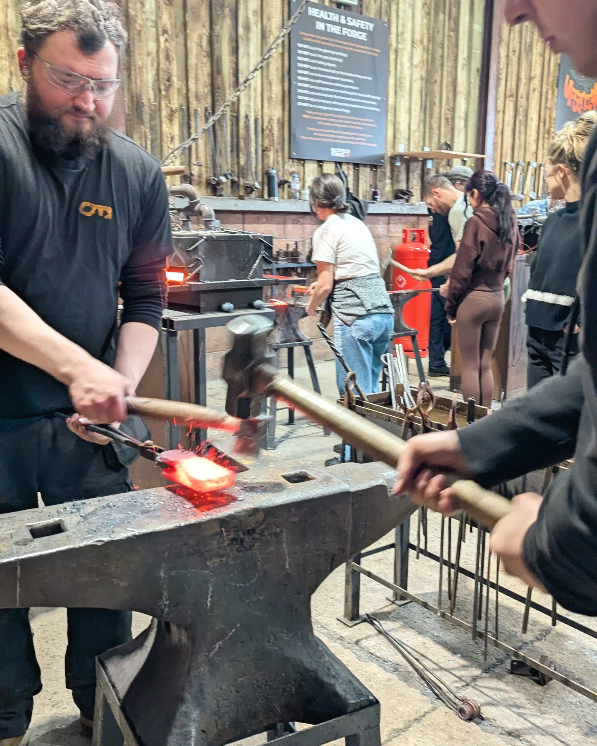 A close-up action shot of a blacksmith hammering the front edge of a glowing orange axe head on the anvil to narrow the profile and shape the blade's taper at Oldfield Forge.