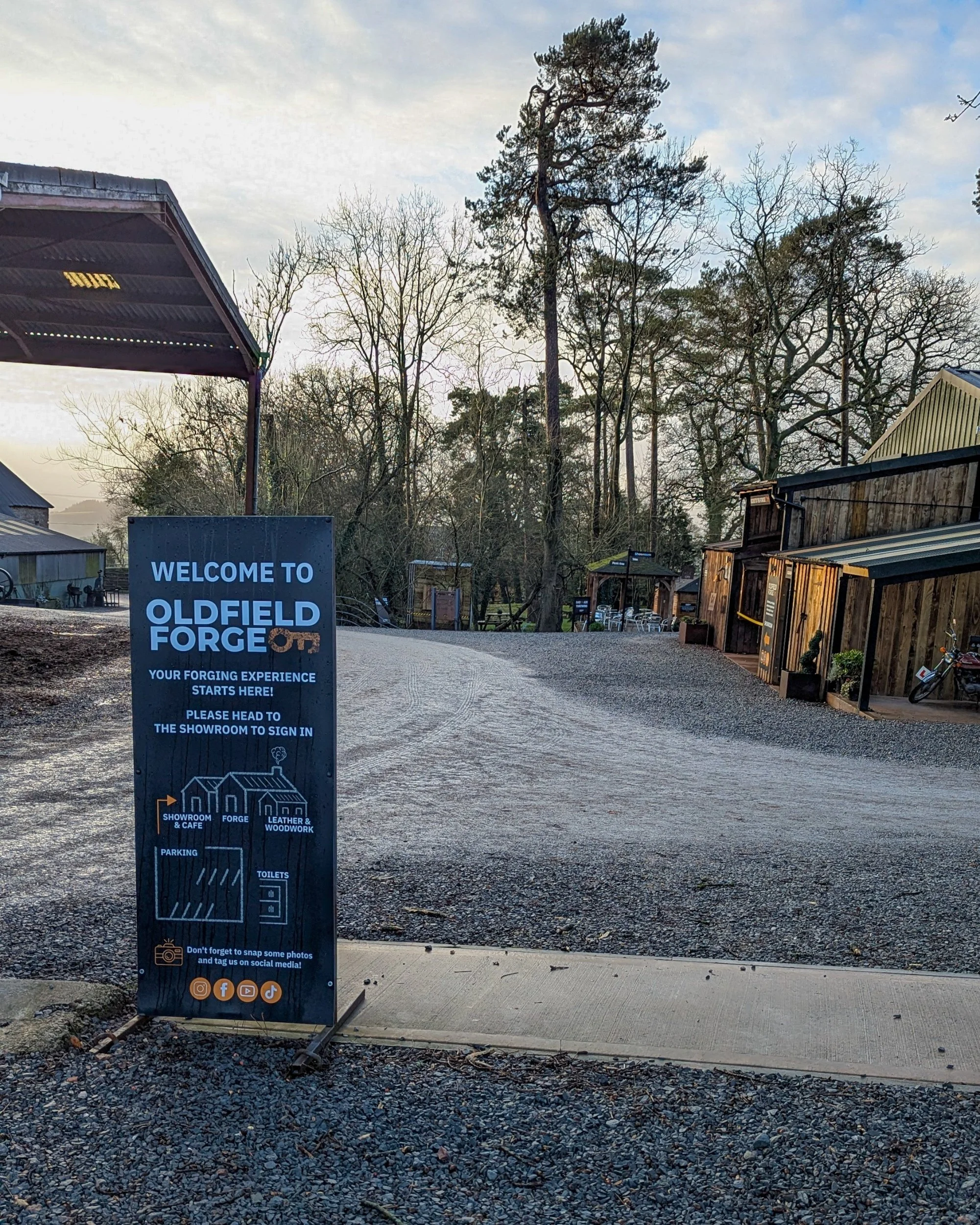 The entrance to Oldfield Forge Barns featuring a rustic wooden sign and a gravel driveway leading toward traditional black-timbered workshop buildings under a cloudy sky.